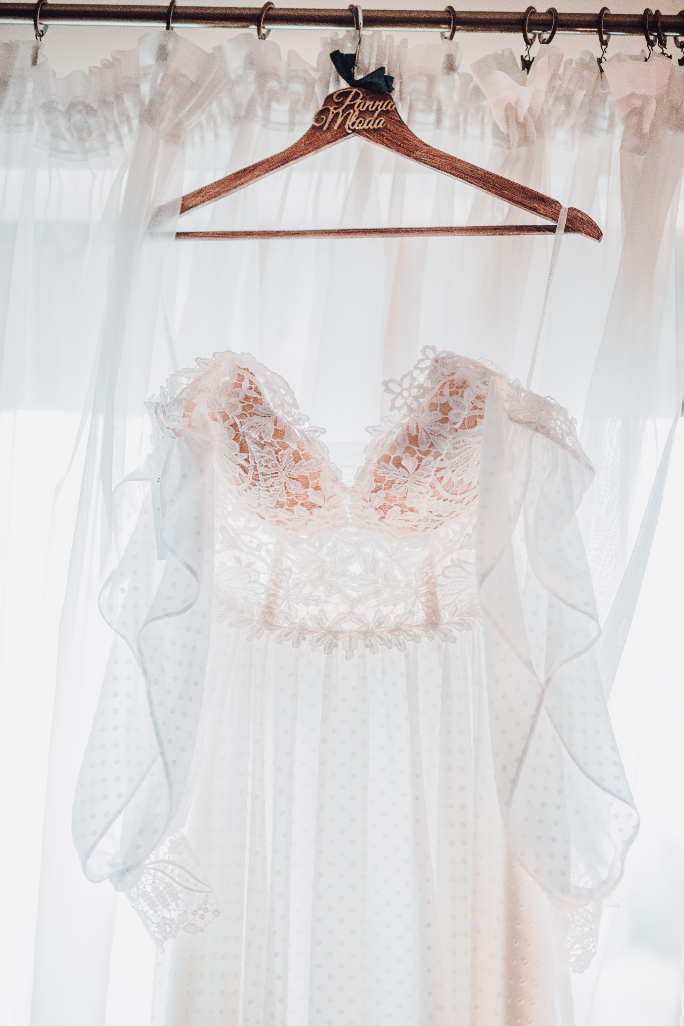 Delikatna suknia ślubna z koronkowym gorsetem i zwiewnym, kropkowanym dołem zawieszona na drewnianym wieszaku z napisem 'Panna Młoda' na tle białych, prześwitujących firan.