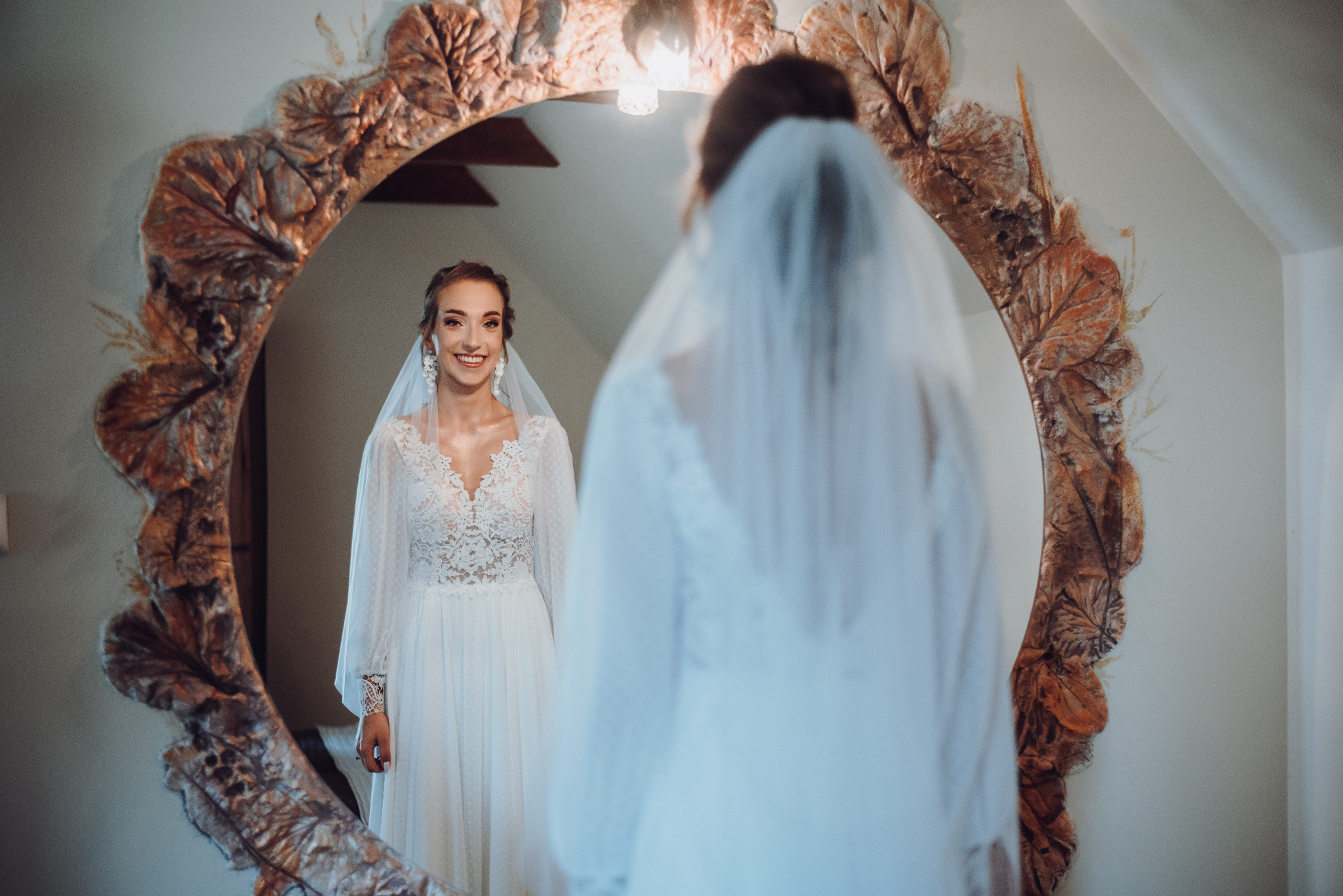 Uśmiechnięta panna młoda w koronkowej sukni z welonem, widziana w ozdobnym lustrze z motywem liści.