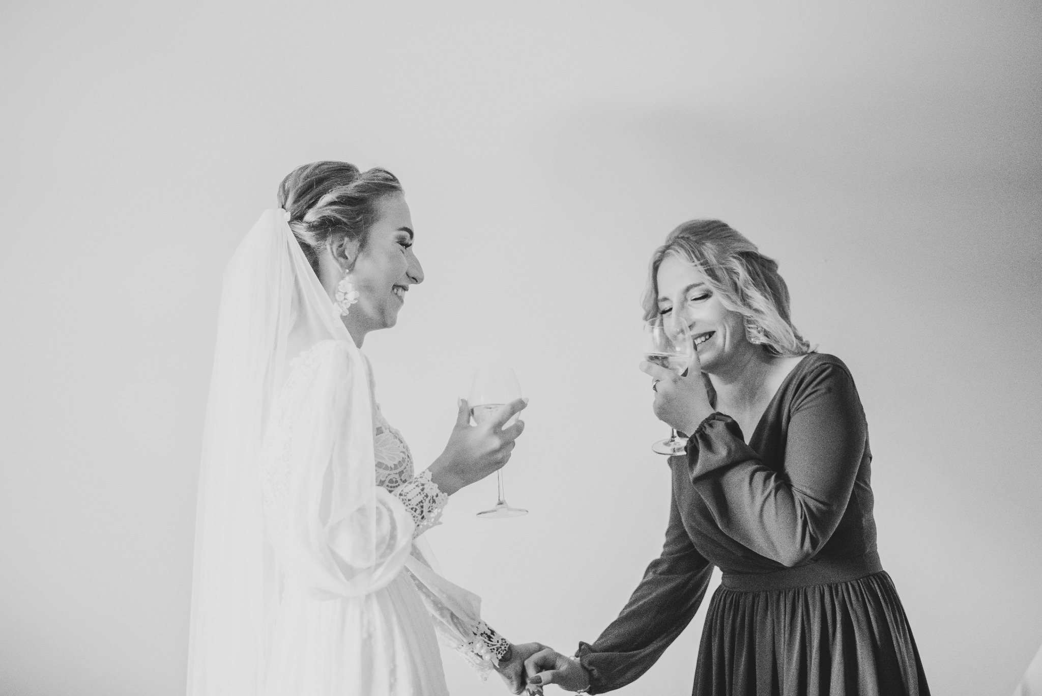 Czarno-białe zdjęcie uśmiechniętej panny młodej w koronkowej sukni i welonie, trzymającej kieliszek z napojem, wznoszącej toast z druhną w sukience z długimi rękawami, trzymając się za ręce.