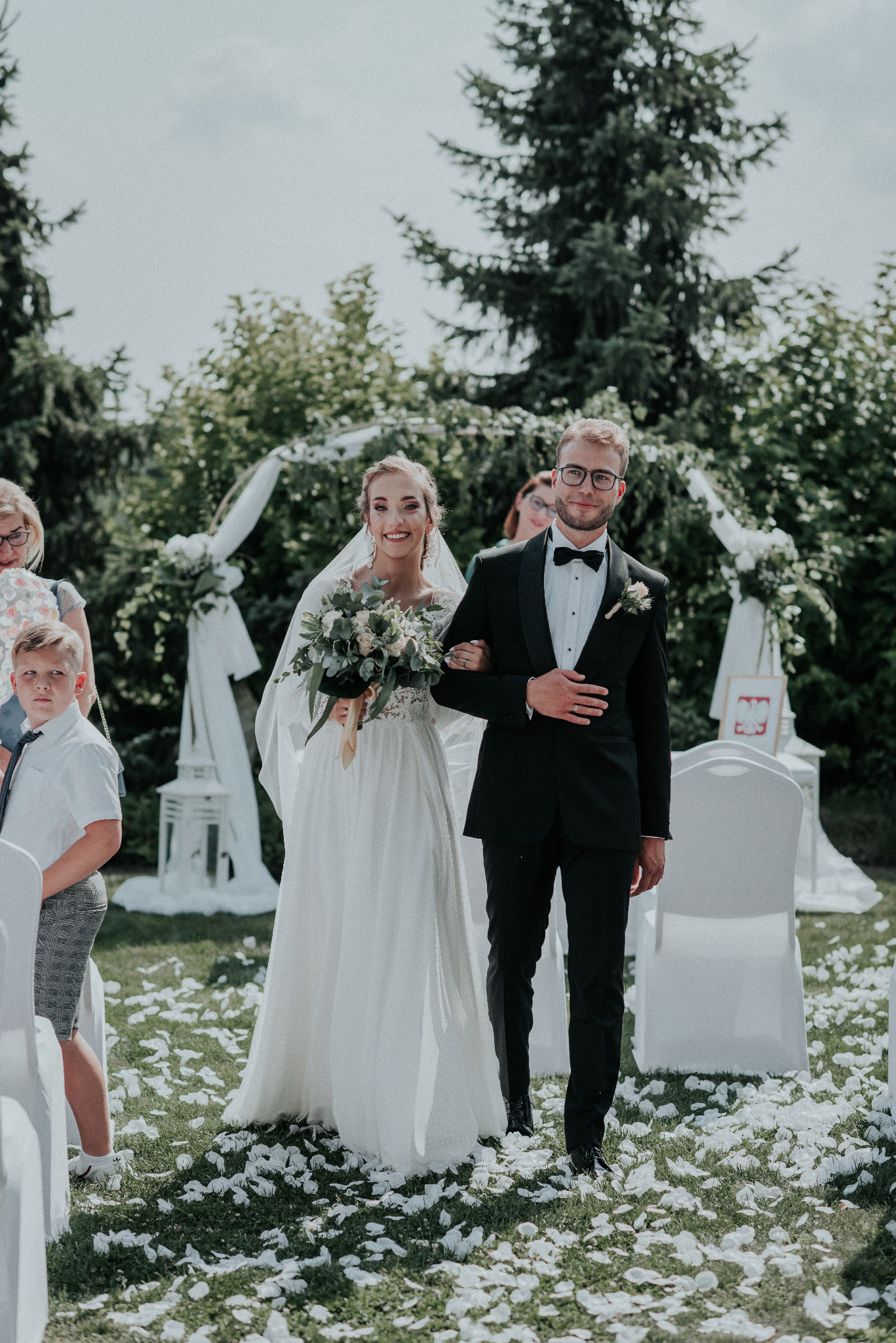 Para młoda w trakcie ceremonii ślubnej na świeżym powietrzu, idą po trawie usłanej białymi płatkami kwiatów, w tle biała dekoracja ślubna i goście.