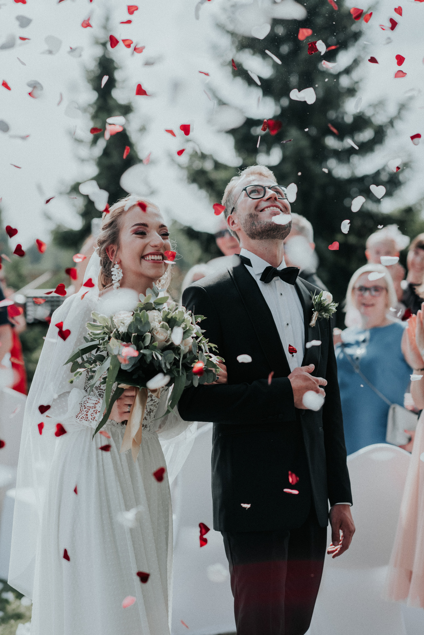Uśmiechnięta para młoda, panna młoda z bukietem, pan młody w smokingu, obsypywana konfetti w kształcie serc na tle zieleni i gości.