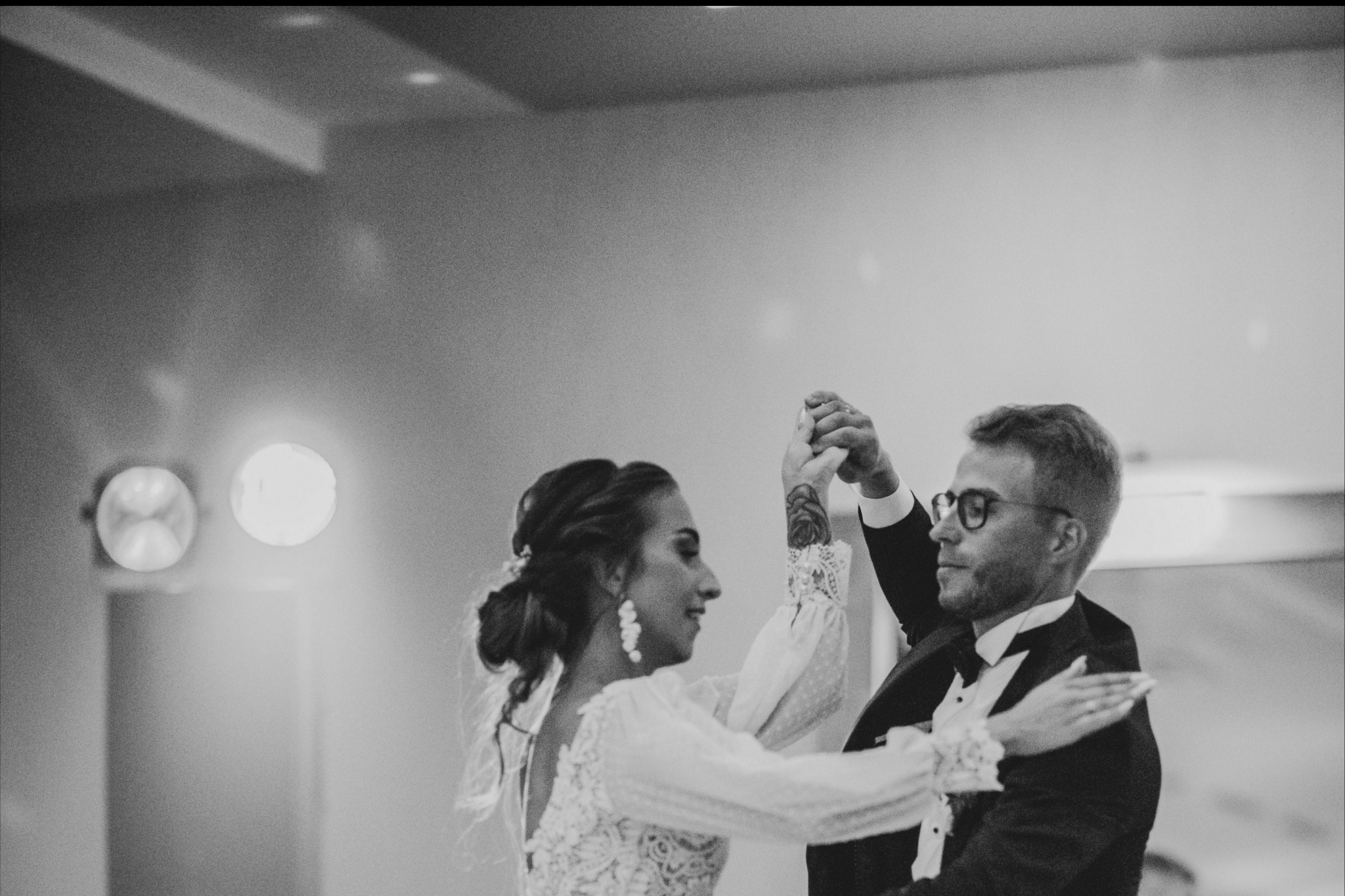 Czarno-białe zdjęcie tańczącej pary: panna młoda w koronkowej sukni z długimi rękawami i tatuażem na ramieniu, pan młody w smokingu i okularach, oboje uśmiechnięci.