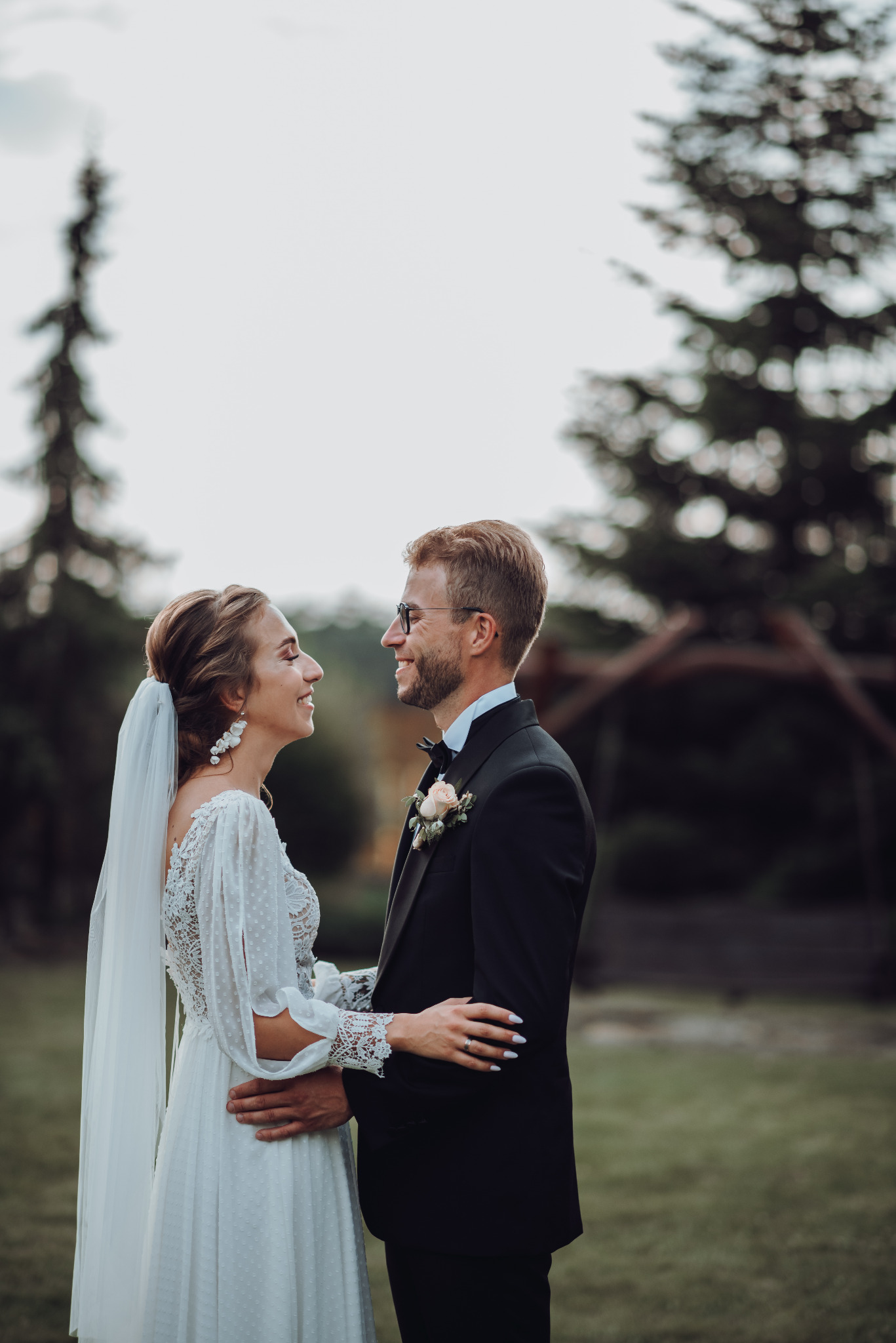 Uśmiechnięta para młoda w plenerze: on w czarnym smokingu z butonierką, ona w białej sukni z koronkowymi rękawami i welonem, patrząca na siebie z czułością.