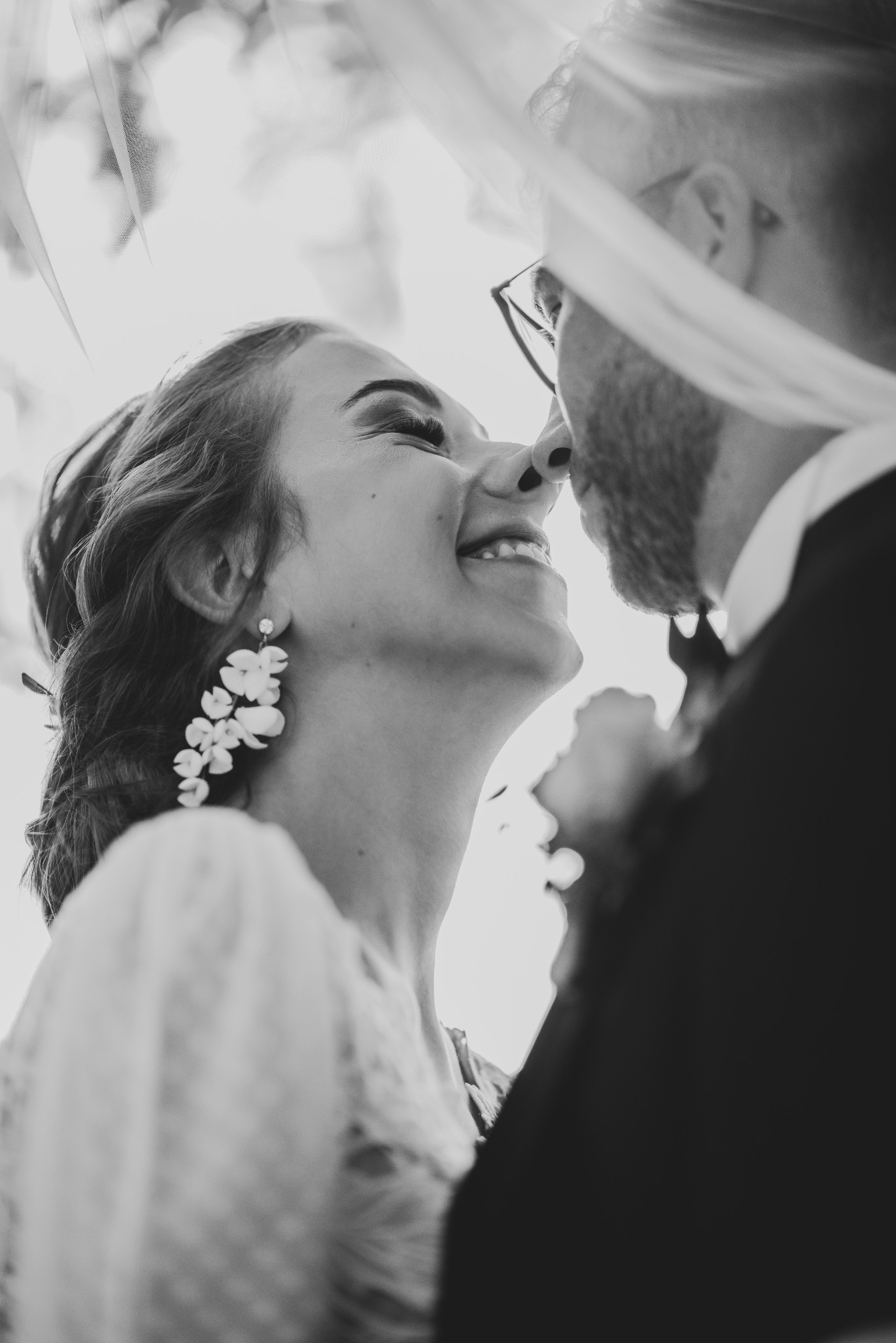 Czarno-białe zbliżenie narzeczonych pod welonem, panna młoda uśmiechnięta, z kwiatowymi kolczykami, pan młody w okularach i garniturze.