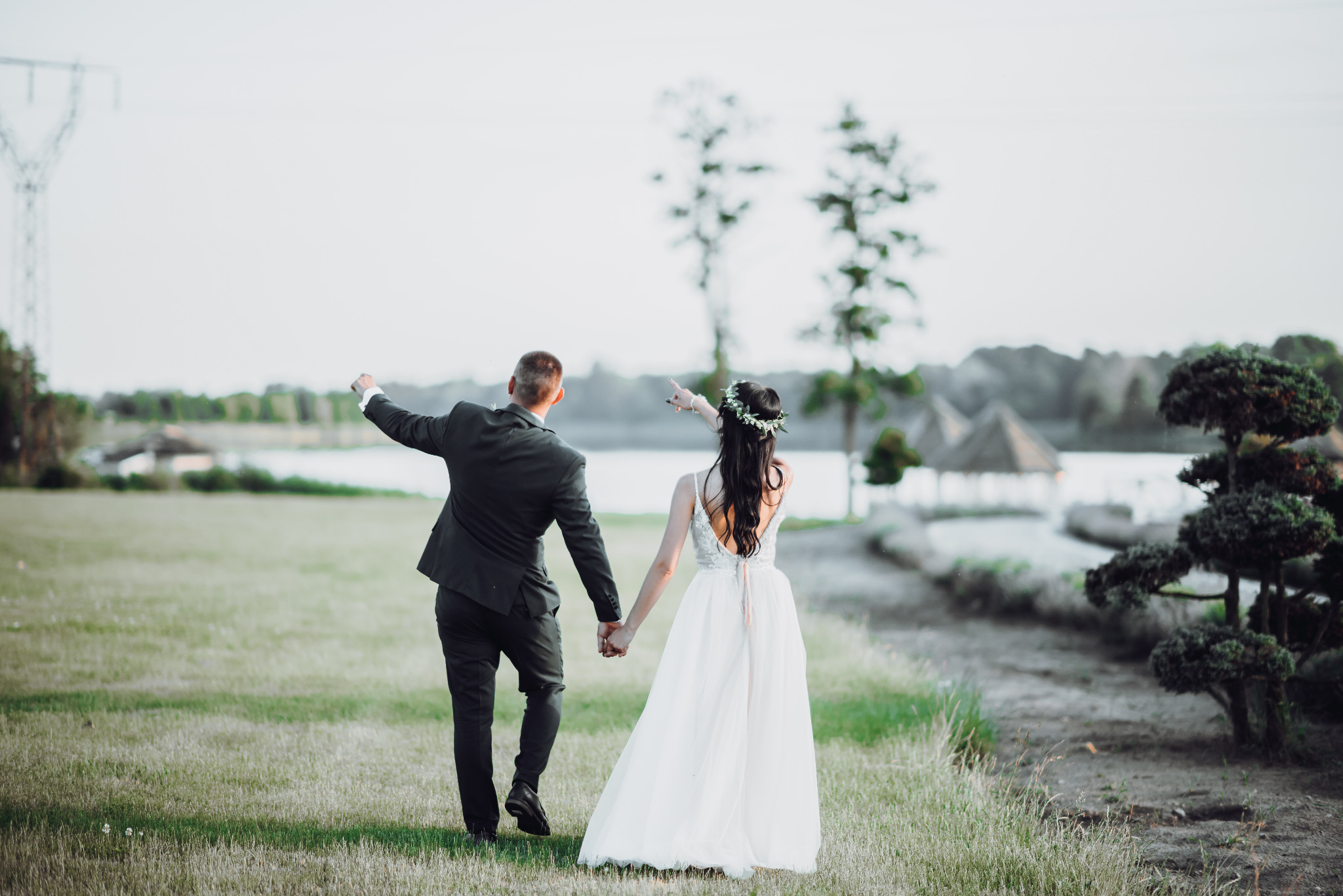 Para młoda, trzymając się za ręce, idzie przez łąkę w kierunku jeziora, on w czarnym garniturze z uniesioną ręką, ona w białej sukni z wiankiem na głowie, widok z tyłu.