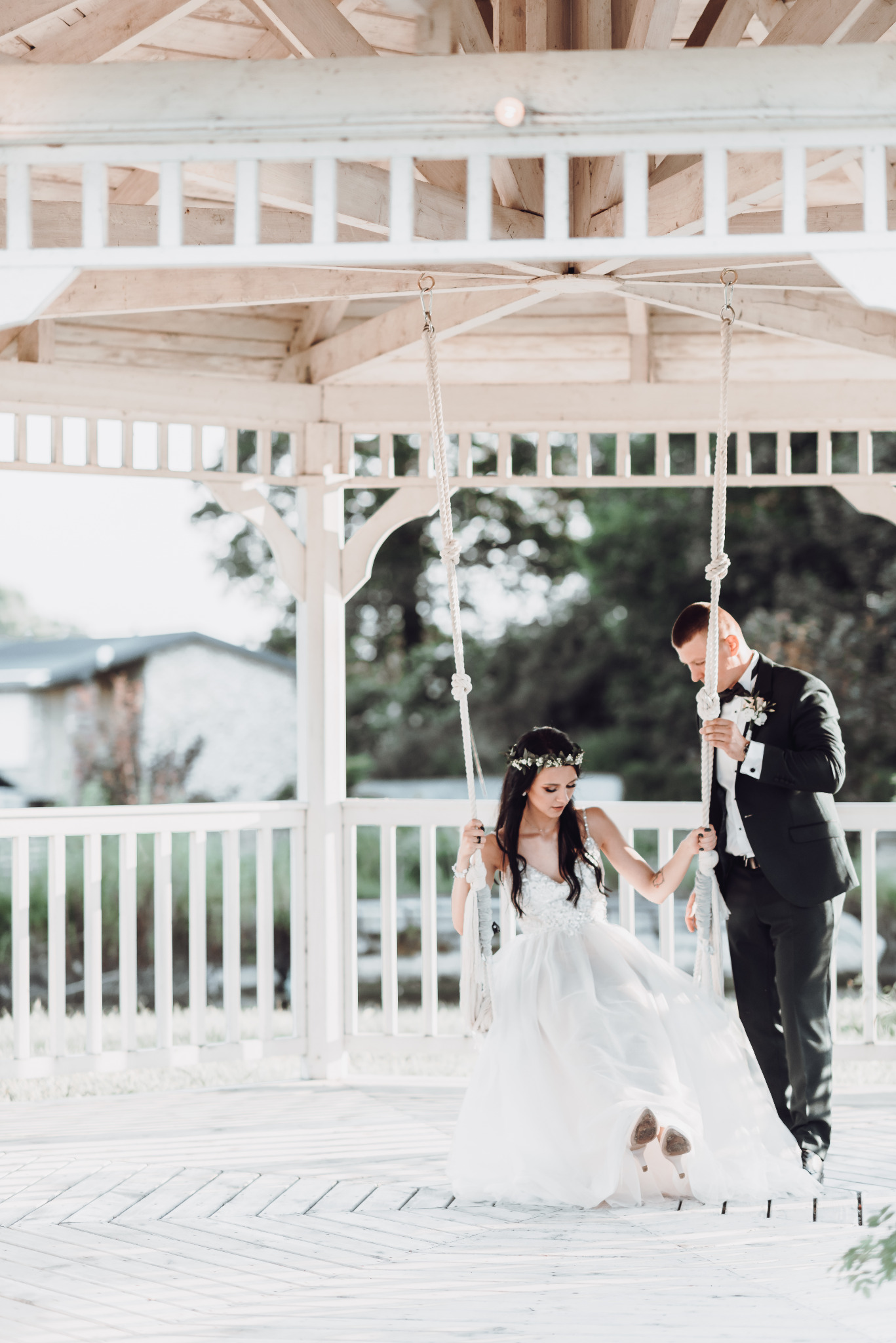 Para młoda w strojach ślubnych na huśtawce w altanie; panna młoda z wiankiem na głowie i w butach na obcasie, pan młody w eleganckim garniturze z muchą.