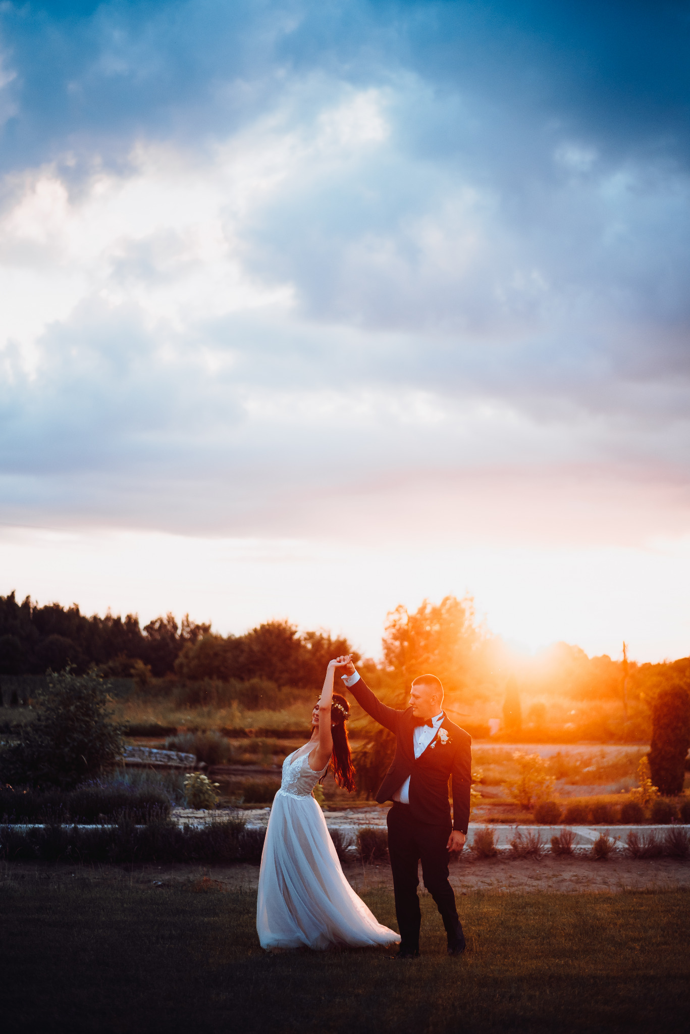 Para w strojach ślubnych tańczy na tle zachodzącego słońca w plenerze, ona w białej sukni z koronką, on w czarnym smokingu z muszką.