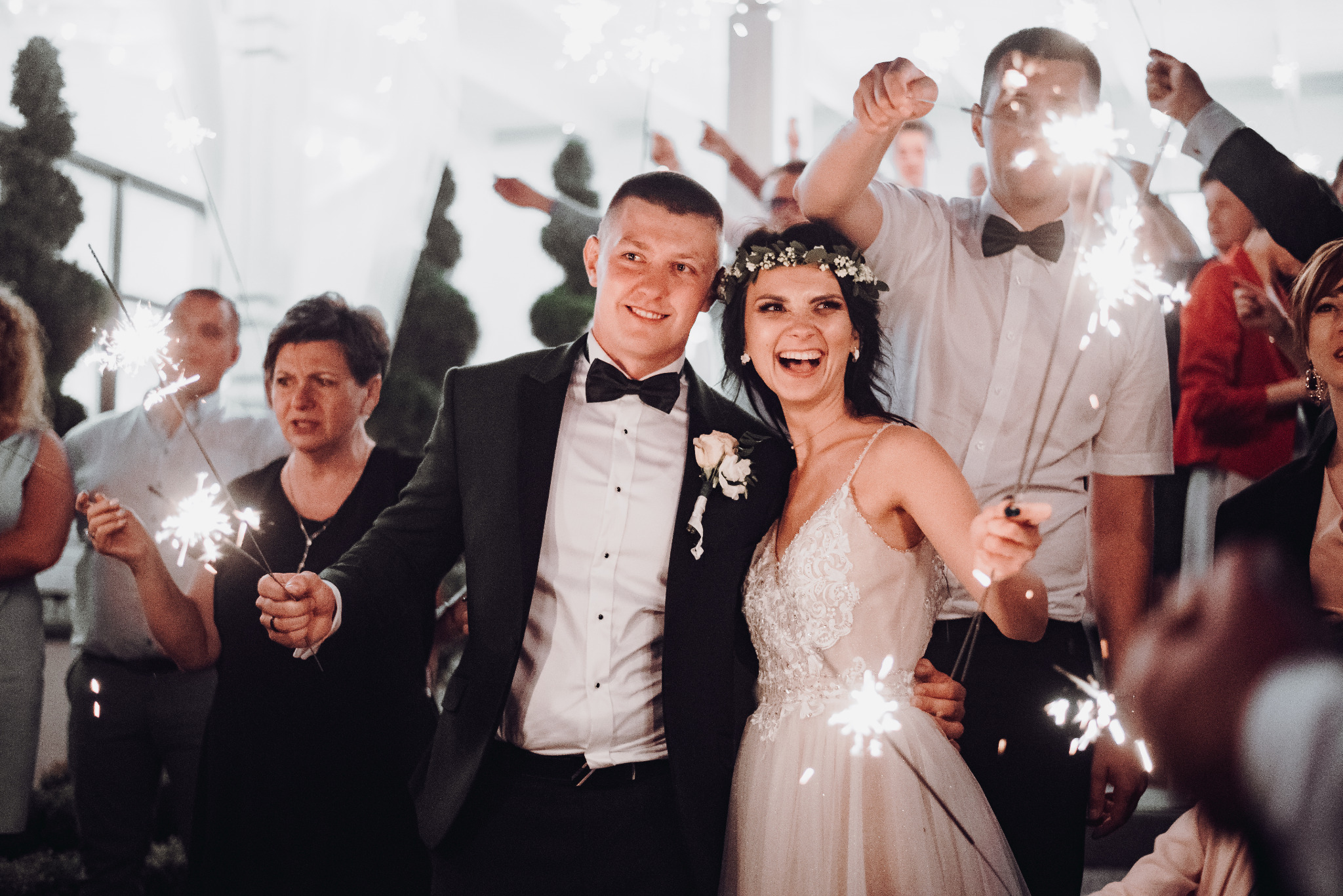 Uśmiechnięta para młoda w otoczeniu gości weselnych trzymających płonące zimne ognie, panna młoda ma wianek na głowie.