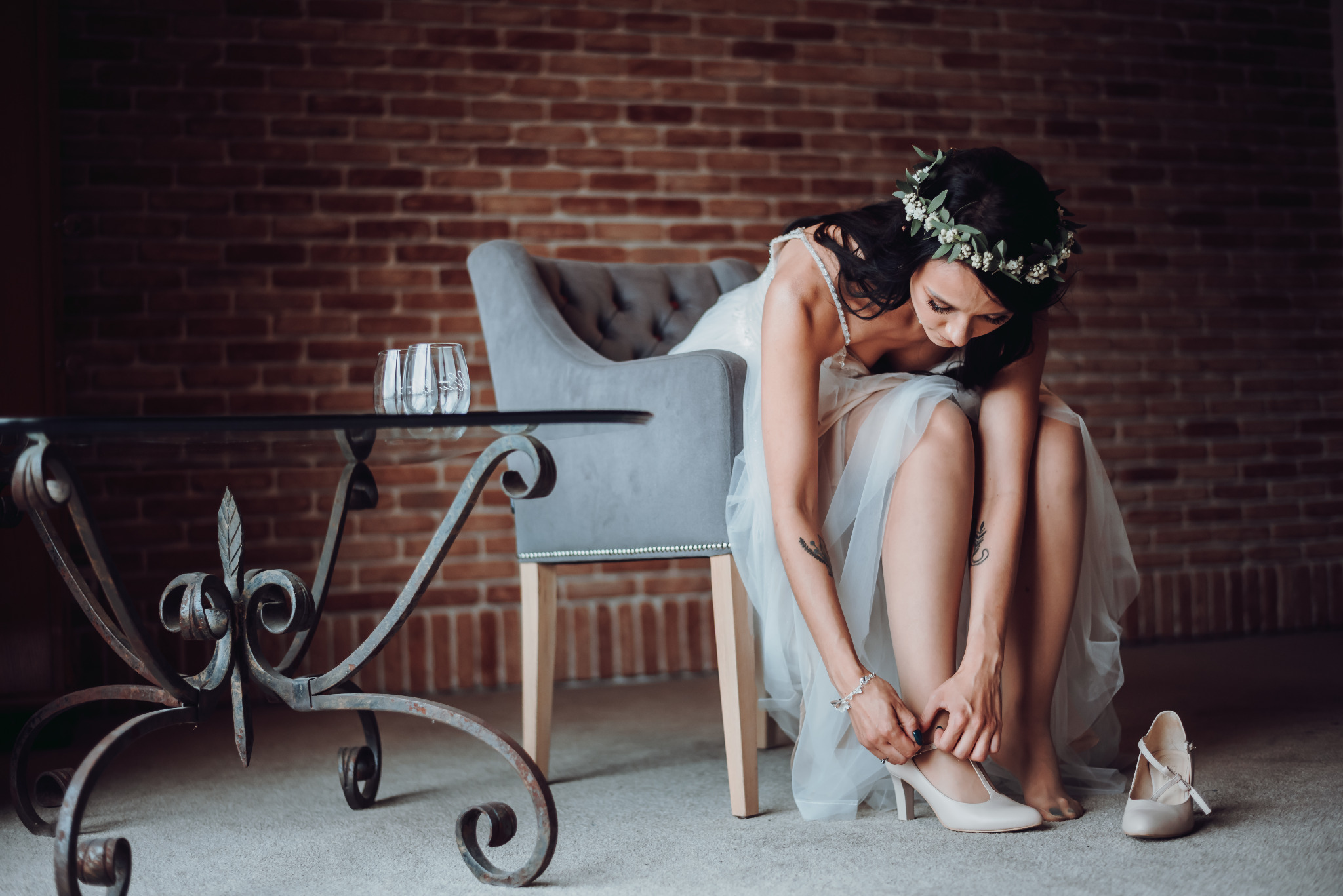 Panna młoda w białej sukni i wianku na głowie, siedząca na szarym krześle, zapina pasek beżowego buta na tle ceglanej ściany.