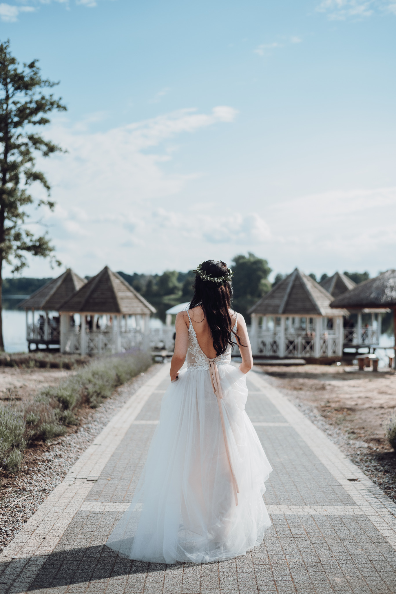 Panna młoda w białej sukni z odkrytymi plecami i wiankiem na głowie, idzie kamienną ścieżką w kierunku drewnianych altan nad jeziorem w słoneczny dzień.