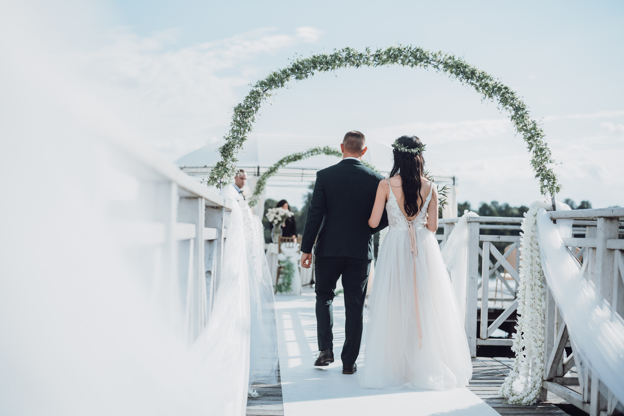 Para młoda, widziana od tyłu, przechodzi przez ozdobny, kwiatowy łuk na drewnianym pomoście w jasny, słoneczny dzień.