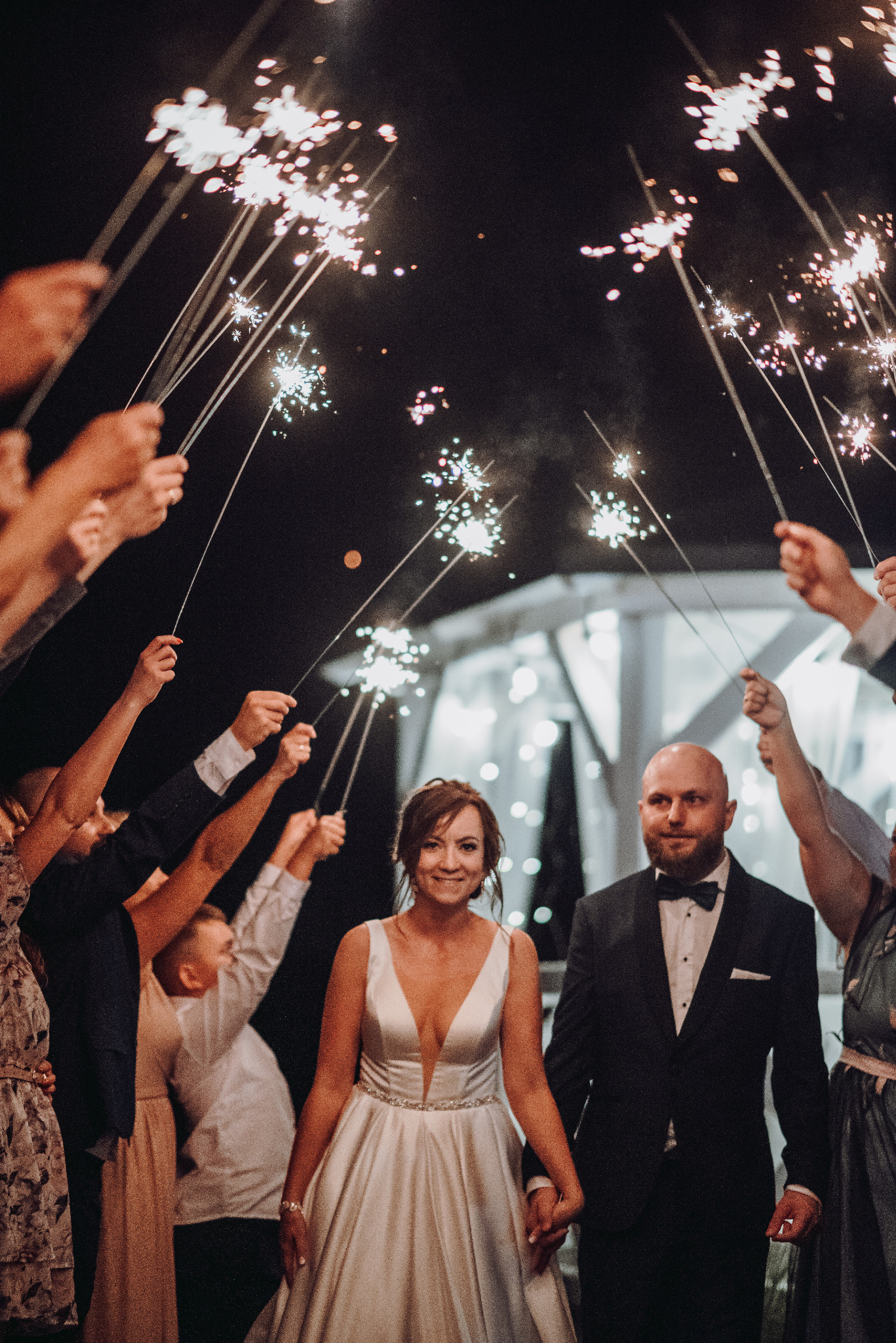 Uśmiechnięta para młoda w eleganckich strojach, trzymająca się za ręce, otoczona przez gości z rozpalonymi zimnymi ogniami podczas wieczornego przyjęcia.