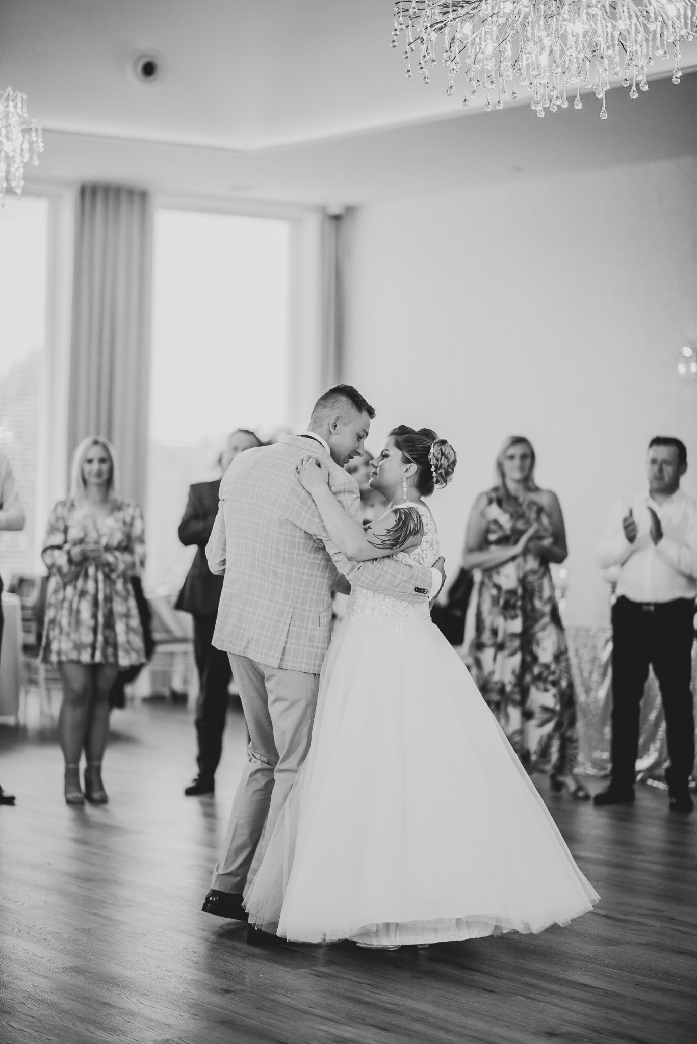 Czarno-białe zdjęcie pary młodej podczas pierwszego tańca, panna młoda w sukni z koronkową górą i upiętymi włosami, pan młody w jasnej marynarce w kratę, w tle goście klaszczący w dłonie.