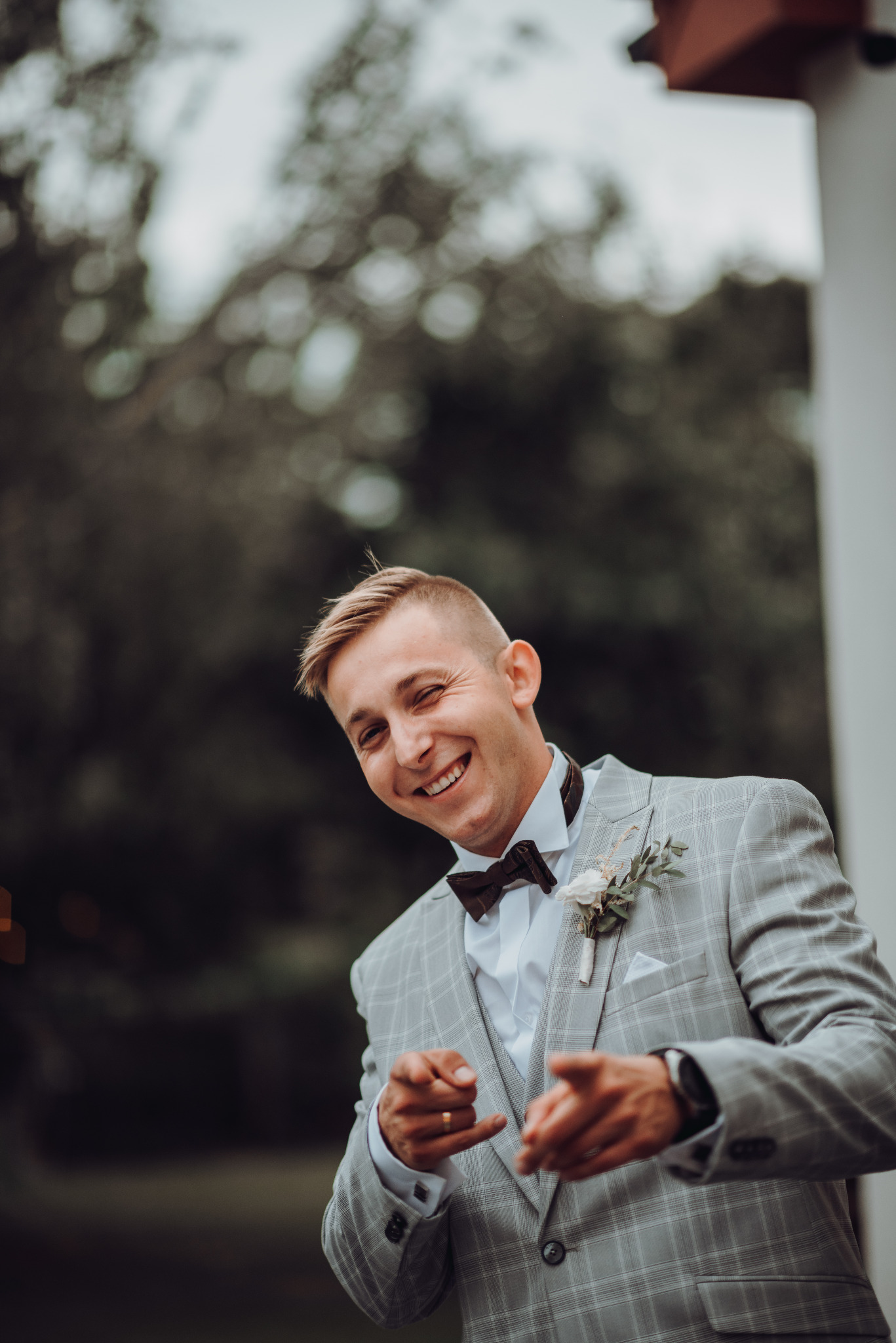 Uśmiechnięty pan młody w szarym garniturze w kratę i z bordową muchą, z butonierką z białych kwiatów, wyciąga dłonie w geście powitania na tle rozmytej zieleni.