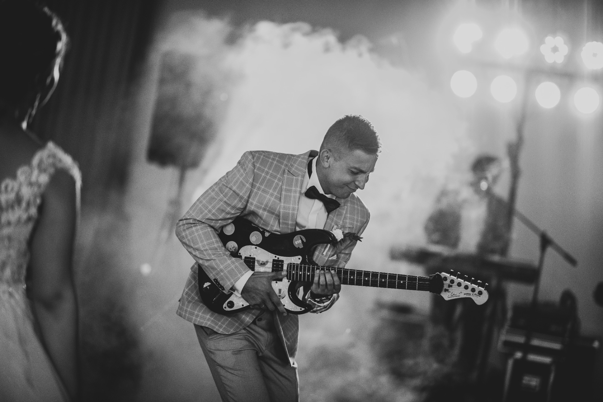 Czarno-białe zdjęcie mężczyzny w kraciastej marynarce i muszce, grającego na czarnej gitarze elektrycznej podczas uroczystości, z rozproszonym oświetleniem i elementami scenografii w tle.