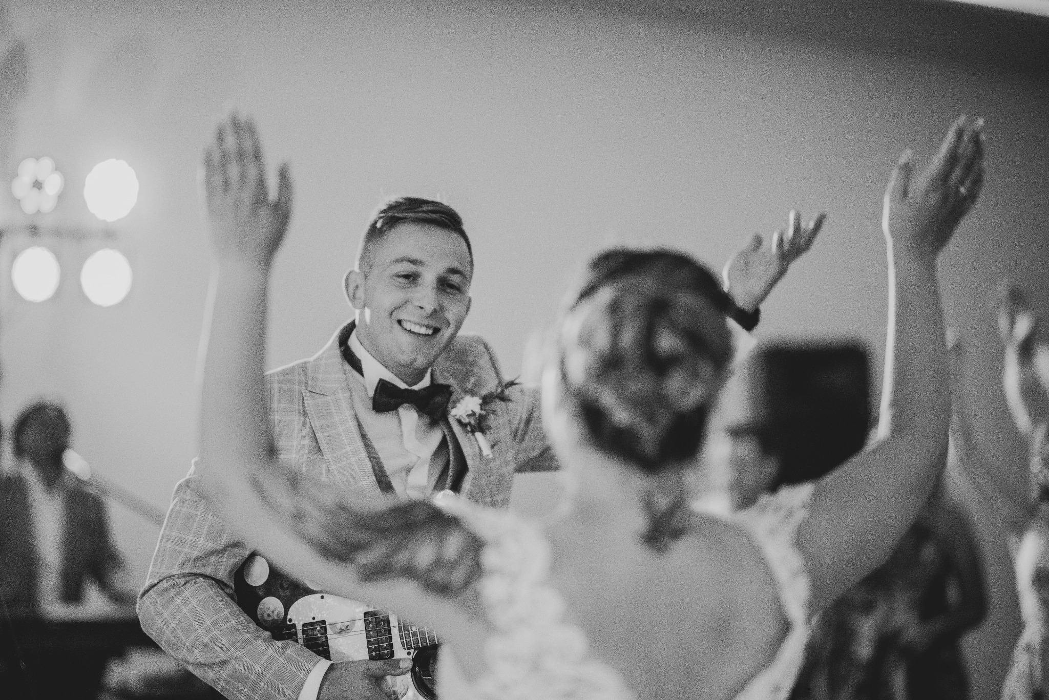 Uśmiechnięty mężczyzna w eleganckiej kraciastej marynarce i muszce gra na gitarze podczas uroczystości, prawdopodobnie wesela, z podniesionymi rękami pary młodej w tle.