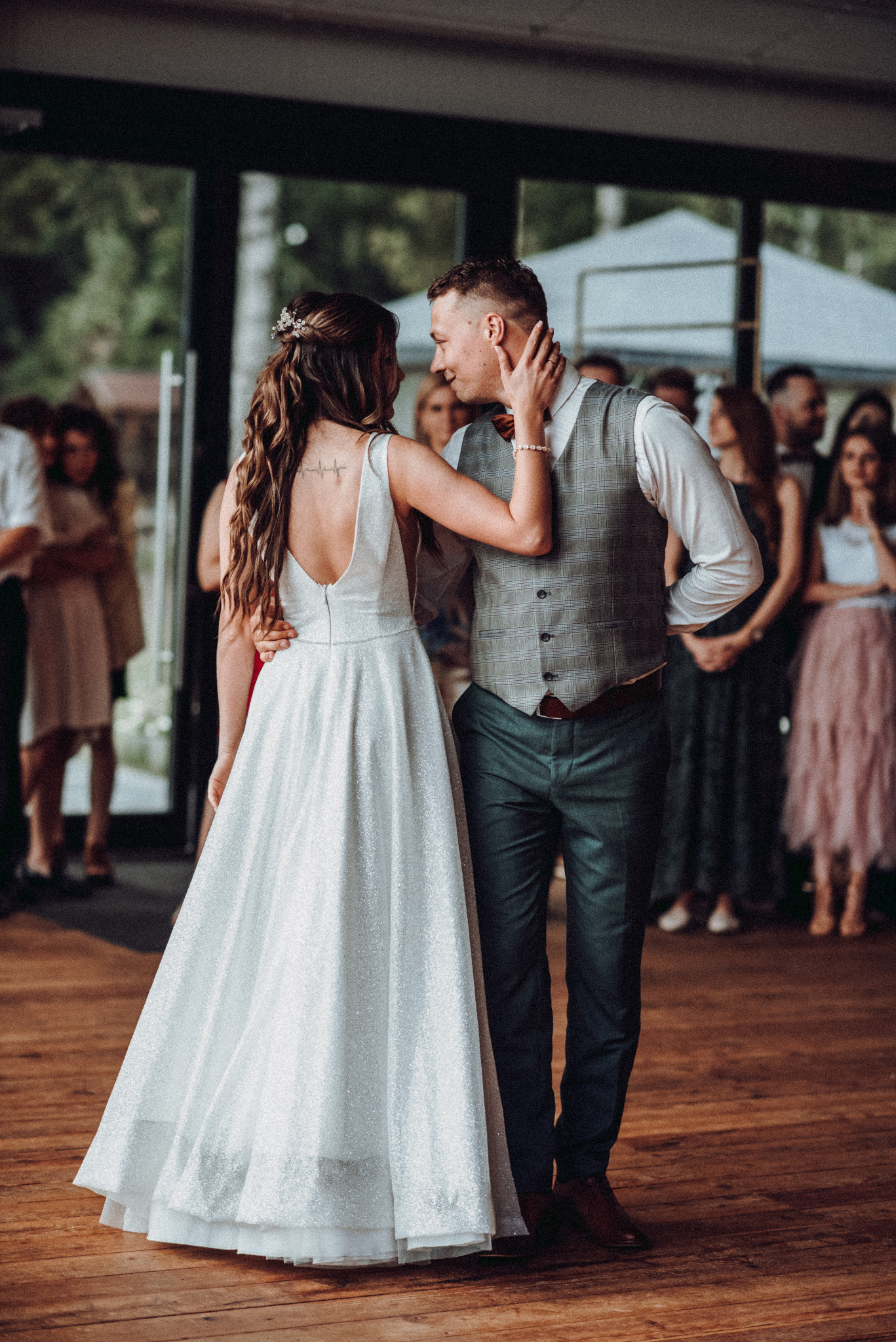 Para młoda w tańcu podczas wesela, panna młoda w białej sukni z odkrytymi plecami i tatuażem, pan młody w kamizelce w kratę i zielonych spodniach, goście weselni w tle.