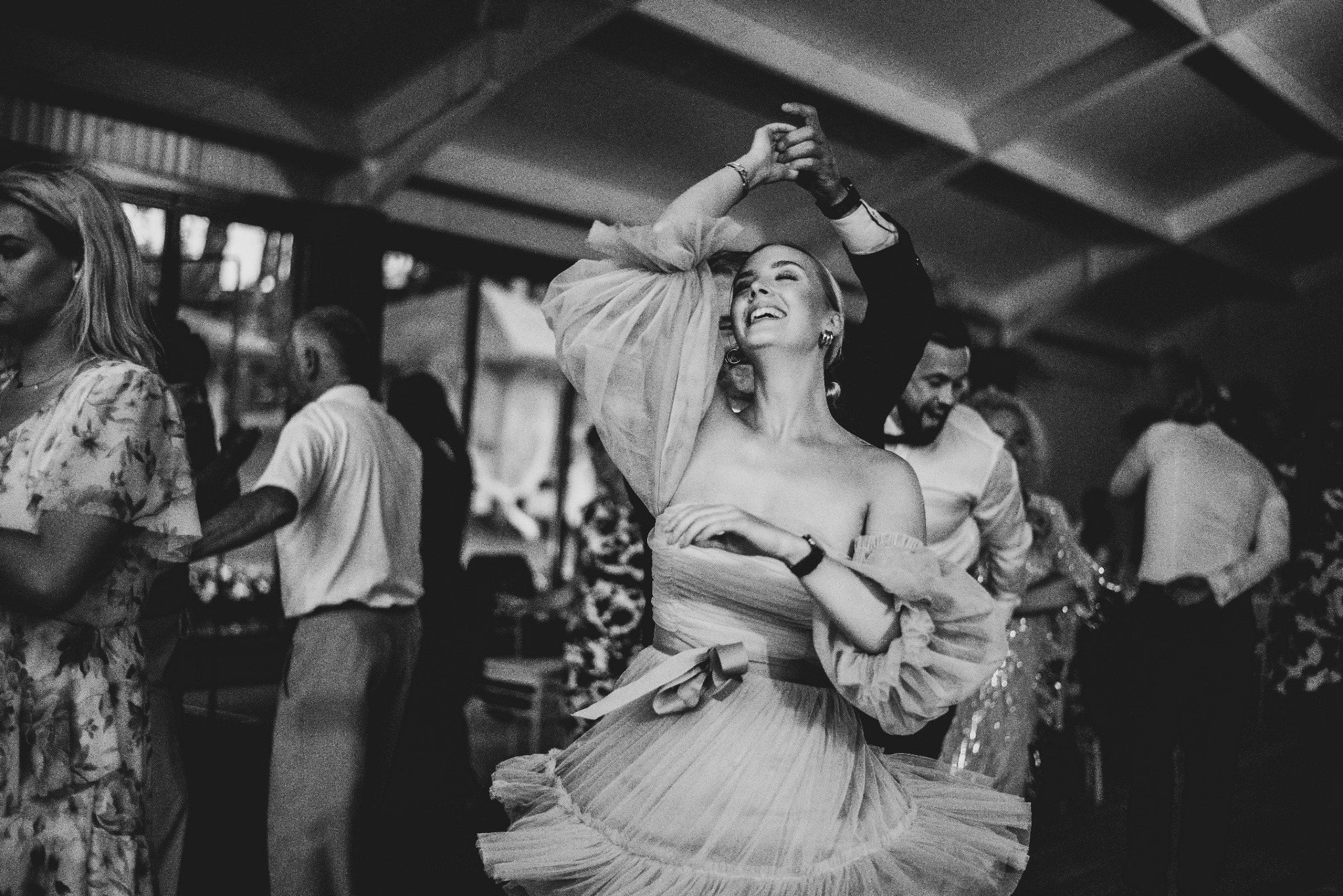 Czarno-białe zdjęcie uśmiechniętej kobiety w tiulowej sukience z bufiastymi rękawami, tańczącej z partnerem na parkiecie podczas wydarzenia.