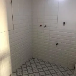 Świeżo wykafelkowana ściana białymi, wypukłymi płytkami typu metro i podłoga z szaro-białymi płytkami w geometryczny wzór w małej łazience. Widoczne wystające rury instalacji wodnej i fragment...