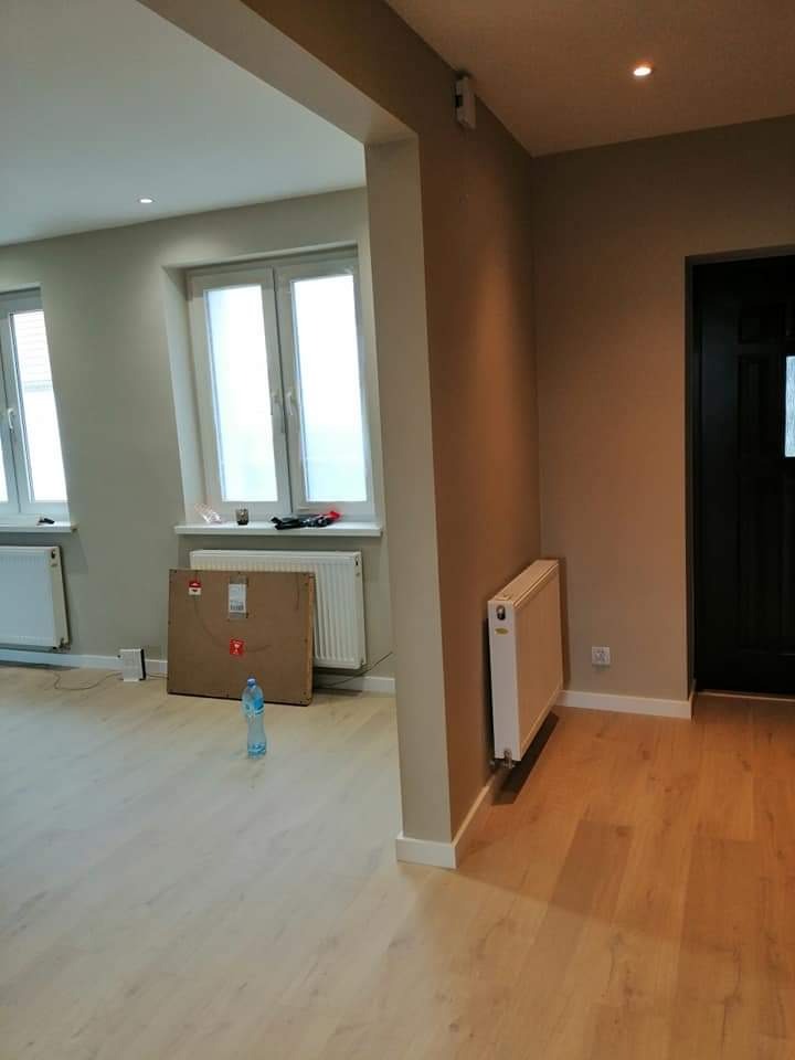 Wnętrze po remoncie: świeżo pomalowane ściany w odcieniach beżu i szarości, jasna drewniana podłoga, białe grzejniki, okno z białymi ramami, kartonowa płyta oparta o ścianę, butelka wody na podłodze.