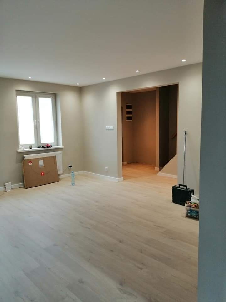 Jasne, świeżo wykończone wnętrze z panelami podłogowymi, widoczne detale jak listwy przypodłogowe, otwór drzwiowy do kolejnego pomieszczenia i kartonowe zabezpieczenie przy grzejniku.