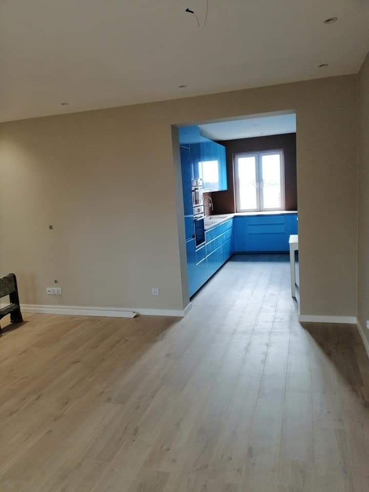 Wnętrze mieszkania po remoncie: jasna drewniana podłoga, beżowe ściany i widok na niebieską, nowoczesną kuchnię z oknem.