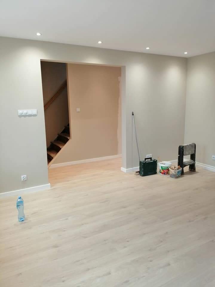 Jasne, nowo wykończone wnętrze z drewnianą podłogą, świeżo pomalowanymi ścianami w odcieniach beżu, widoczne schody w tle i akcesoria malarskie przy ścianie.
