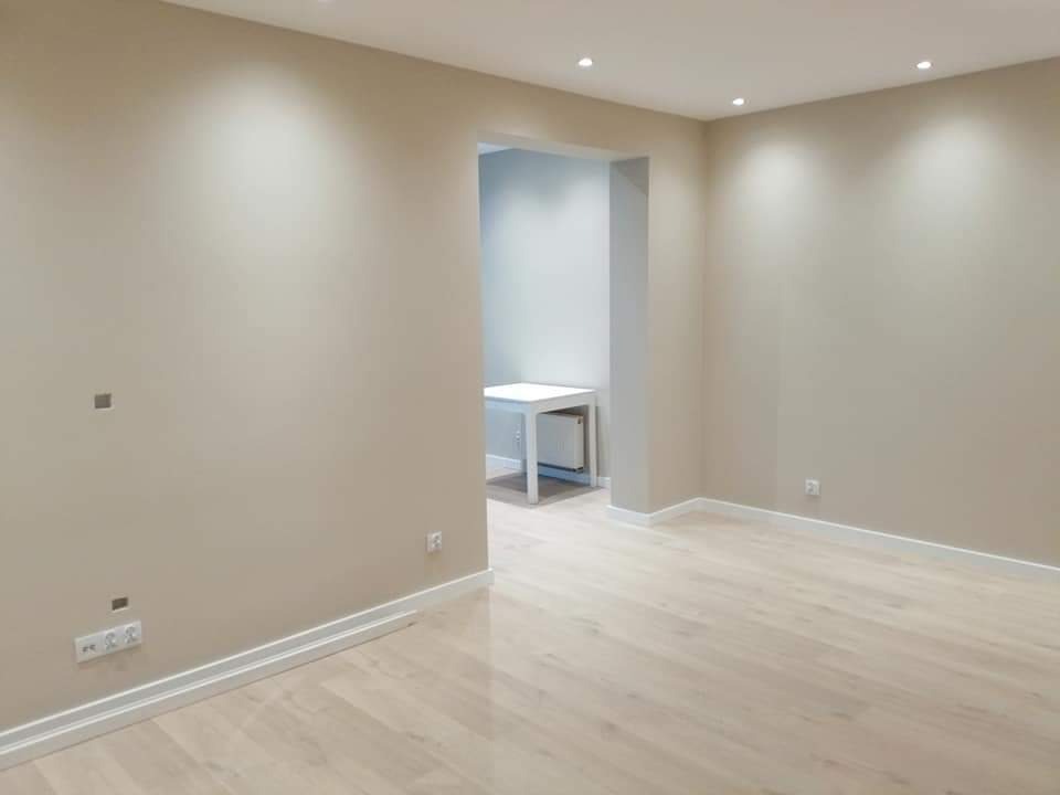 Jasne, świeżo wykończone wnętrze z beżowymi ścianami, białymi listwami przypodłogowymi i panelami podłogowymi w odcieniach bieli i szarości, z widocznymi gniazdkami elektrycznymi i otworami po...