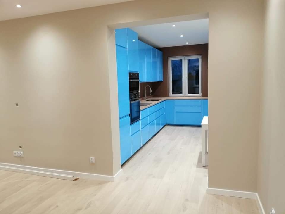 Nowoczesna kuchnia z błękitnymi szafkami, brązową ścianą i jasną podłogą, widziana z sąsiedniego pokoju z beżowymi ścianami.