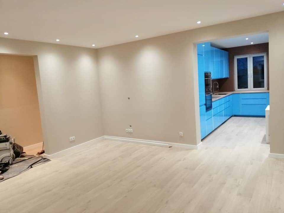 Jasny pokój z nową, jasną podłogą i świeżo pomalowanymi ścianami w odcieniach beżu, z widokiem na kuchnię z nowoczesną, błękitną zabudową.