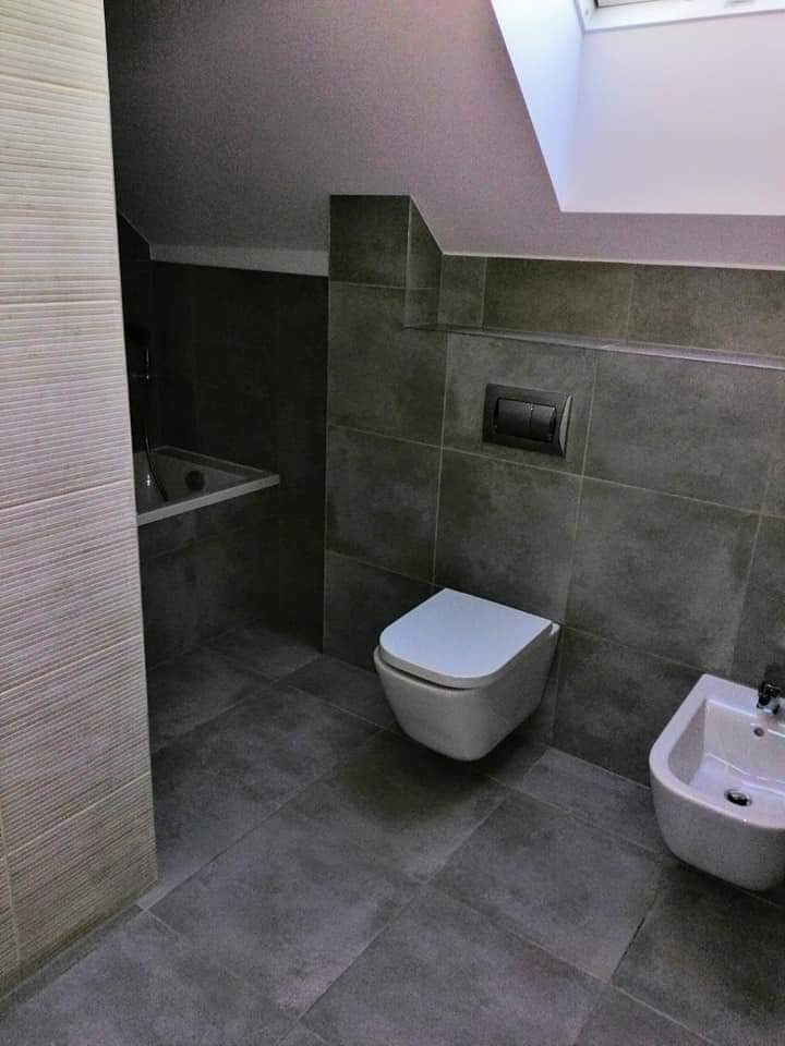 Szara łazienka na poddaszu ze skośnym sufitem i oknem dachowym, wyposażona w wiszącą toaletę, bidet oraz wannę w zabudowie.