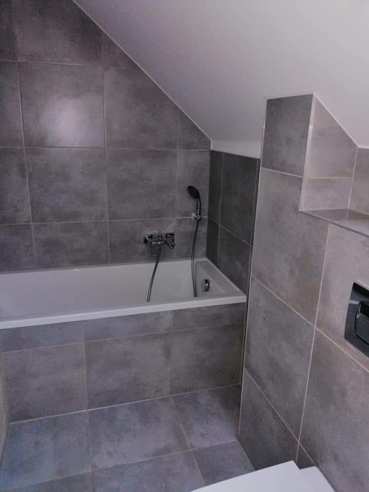 Szara łazienka na poddaszu z wanną obłożoną płytkami w tym samym odcieniu, z widocznym panelem sterującym spłuczką.