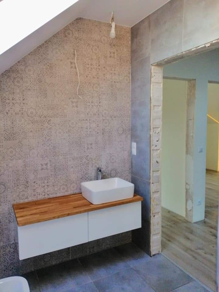 Nowoczesna łazienka na poddaszu z umywalką nablatową na drewnianym blacie, wzorzyste płytki na ścianie pod skosem, surowy otwór drzwiowy i szare płytki podłogowe.