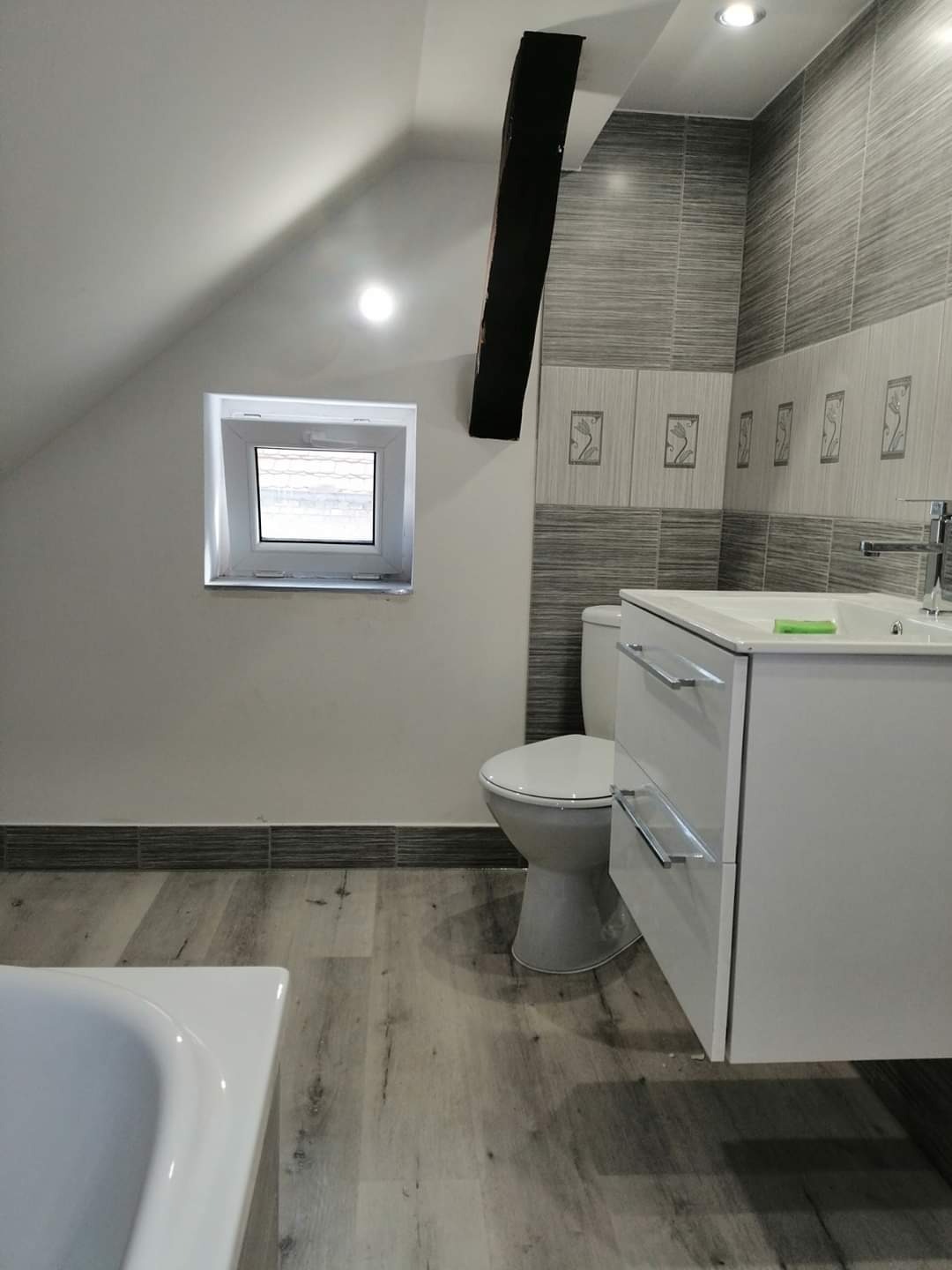 Szara łazienka na poddaszu z oknem dachowym, płytkami imitującymi drewno i białą szafką pod umywalką z chromowanymi uchwytami.