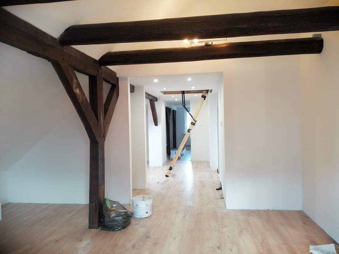 Wnętrze po remoncie z drewnianymi belkami stropowymi, nową podłogą i świeżo pomalowanymi ścianami, z widoczną drabiną i workiem na śmieci.