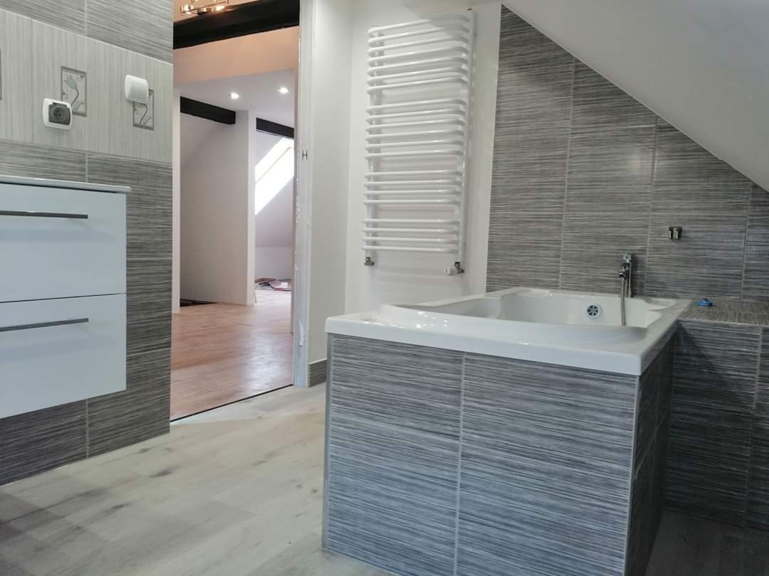 Nowoczesna łazienka z wanną obudowaną szarą, poziomą mozaiką, białą szafką z szufladami i grzejnikiem drabinkowym na ścianie, widok na poddasze przez otwarte drzwi.