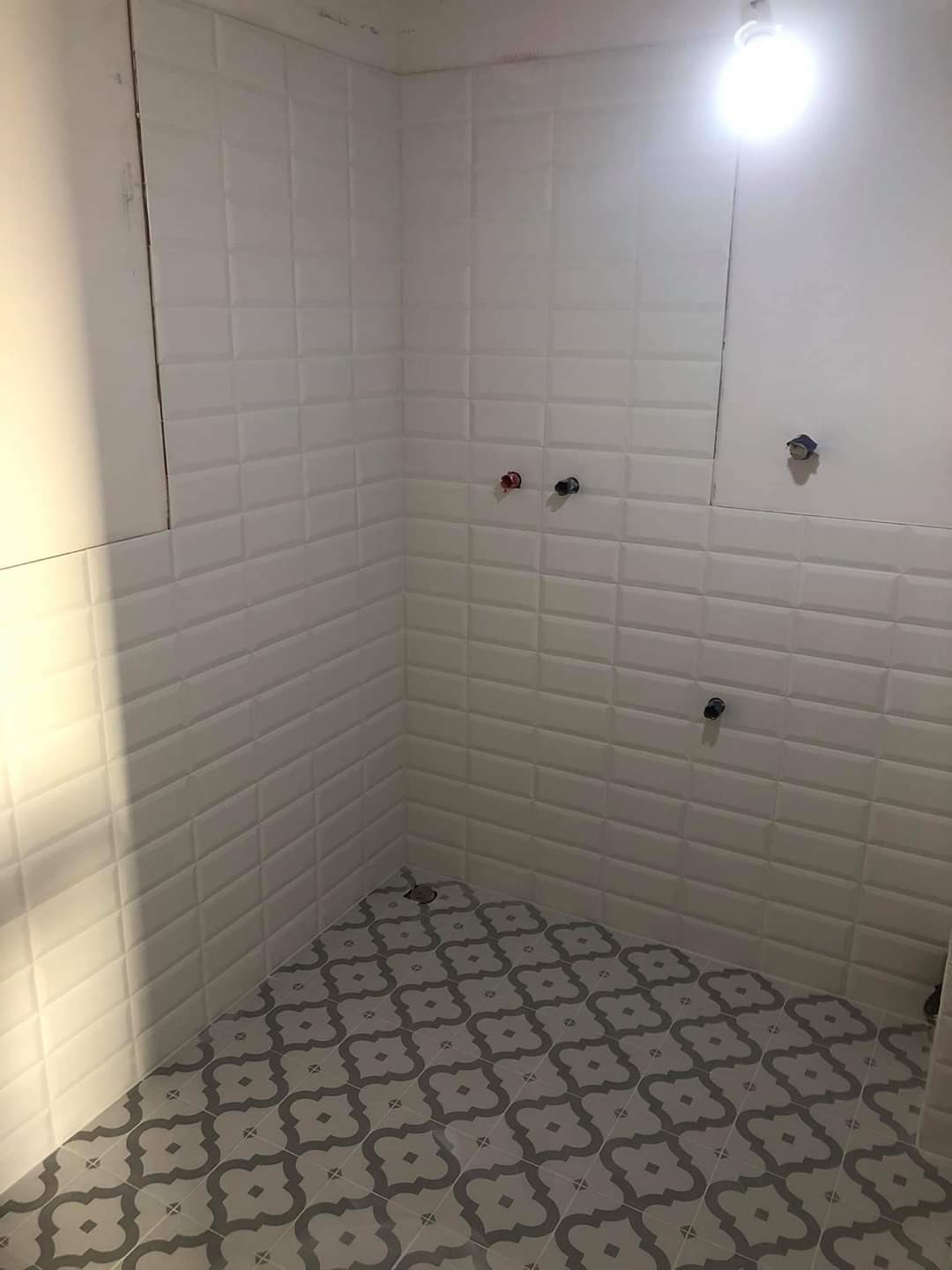 Świeżo wykafelkowana ściana białymi, wypukłymi płytkami typu metro i podłoga z szaro-białymi płytkami w geometryczny wzór w małej łazience. Widoczne wystające rury instalacji wodnej i fragment...