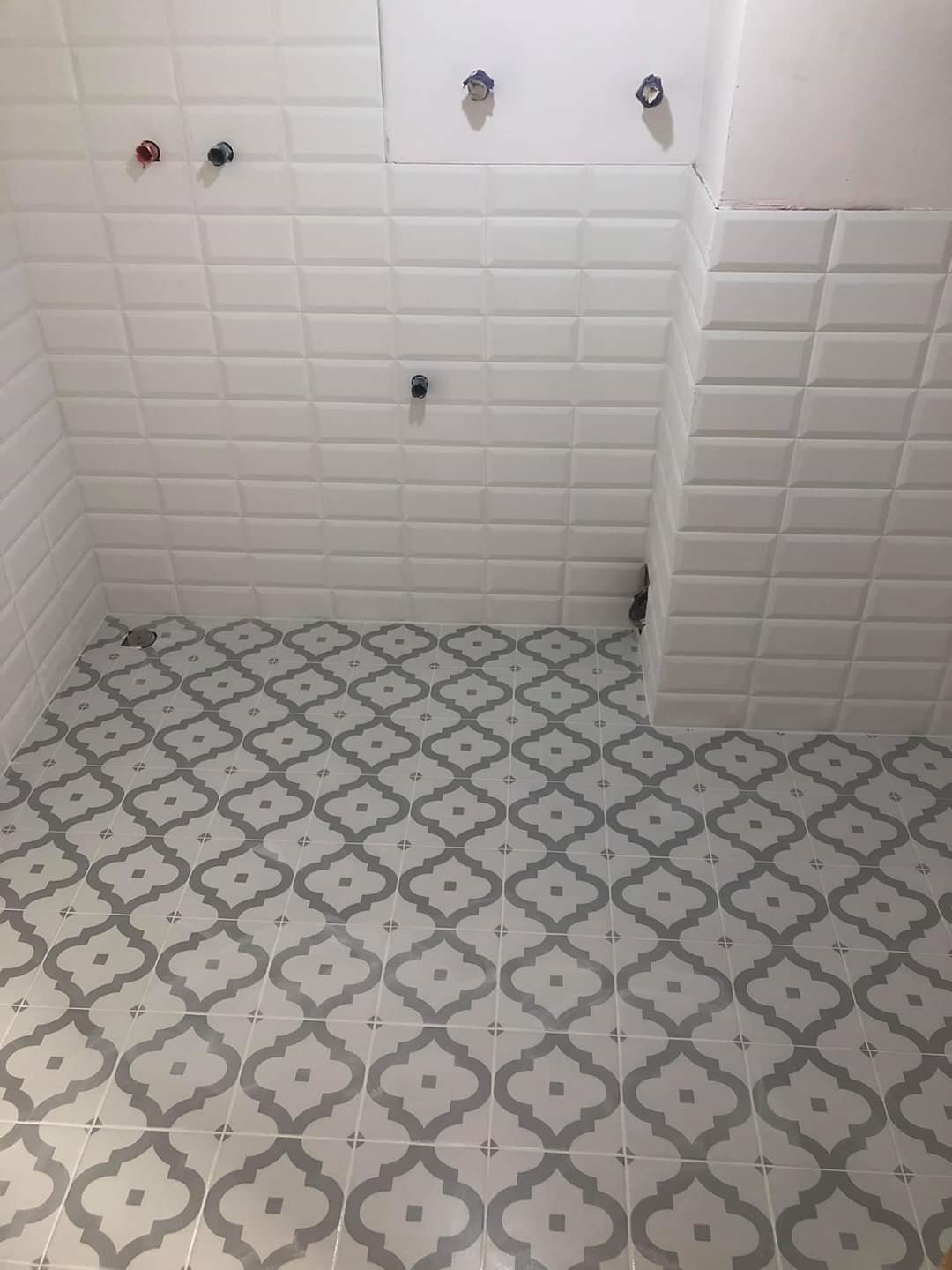 Wykończona łazienka z białymi, wypukłymi płytkami ściennymi w stylu cegiełek i szaro-białą wzorzystą podłogą z płytek. Widoczne instalacje wodne na ścianie.