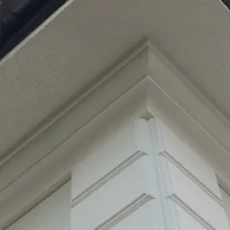 Fragment narożnika budynku po wykonaniu elewacji, widoczne białe zdobienia, szara elewacja, rynna i oświetlenie punktowe.