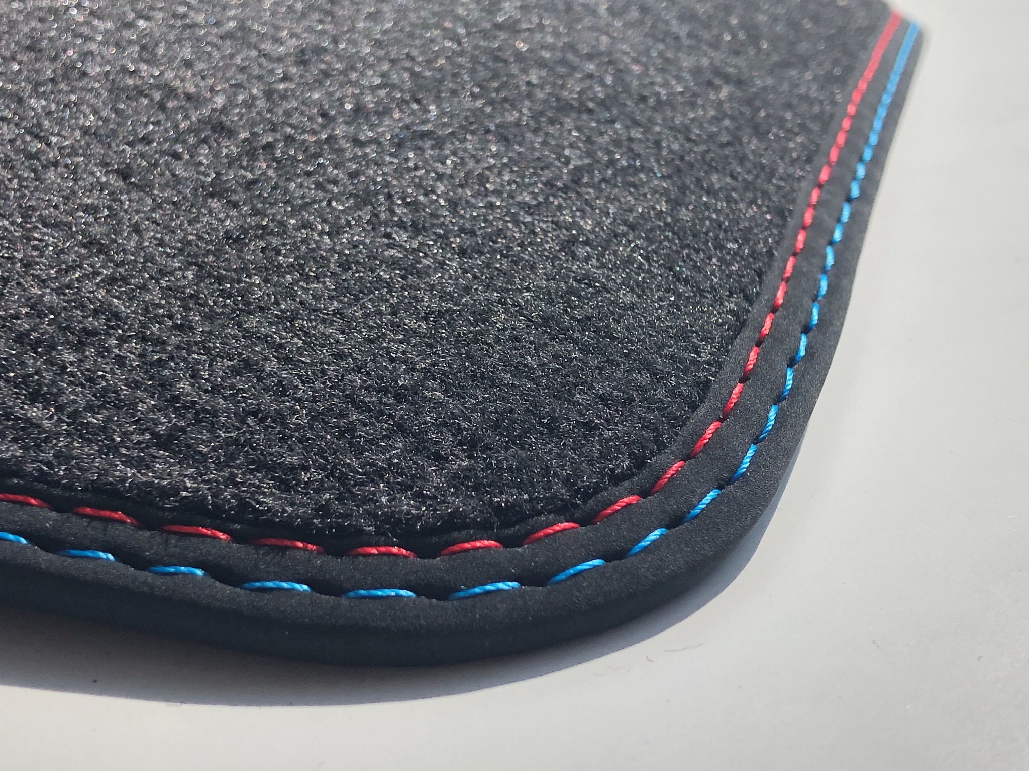 Fragment czarnego dywanika samochodowego z obszyciem w kolorach niebieskim i czerwonym, widoczne z bliska, na jasnym tle.