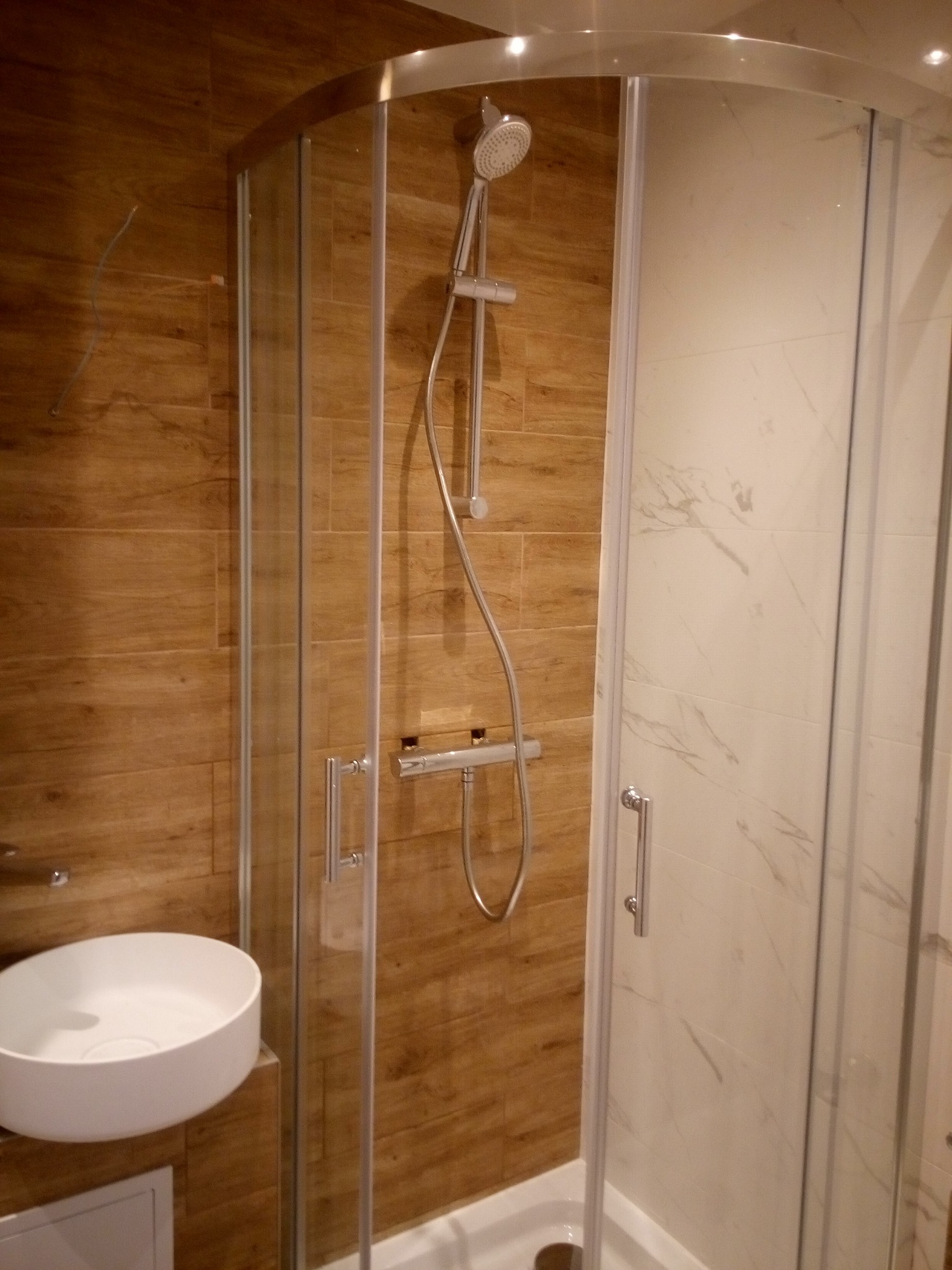 Nowoczesna łazienka z kabiną prysznicową z zaokrąglonymi szybami, białą umywalką nablatową i ścianami wyłożonymi płytkami imitującymi drewno i marmur.