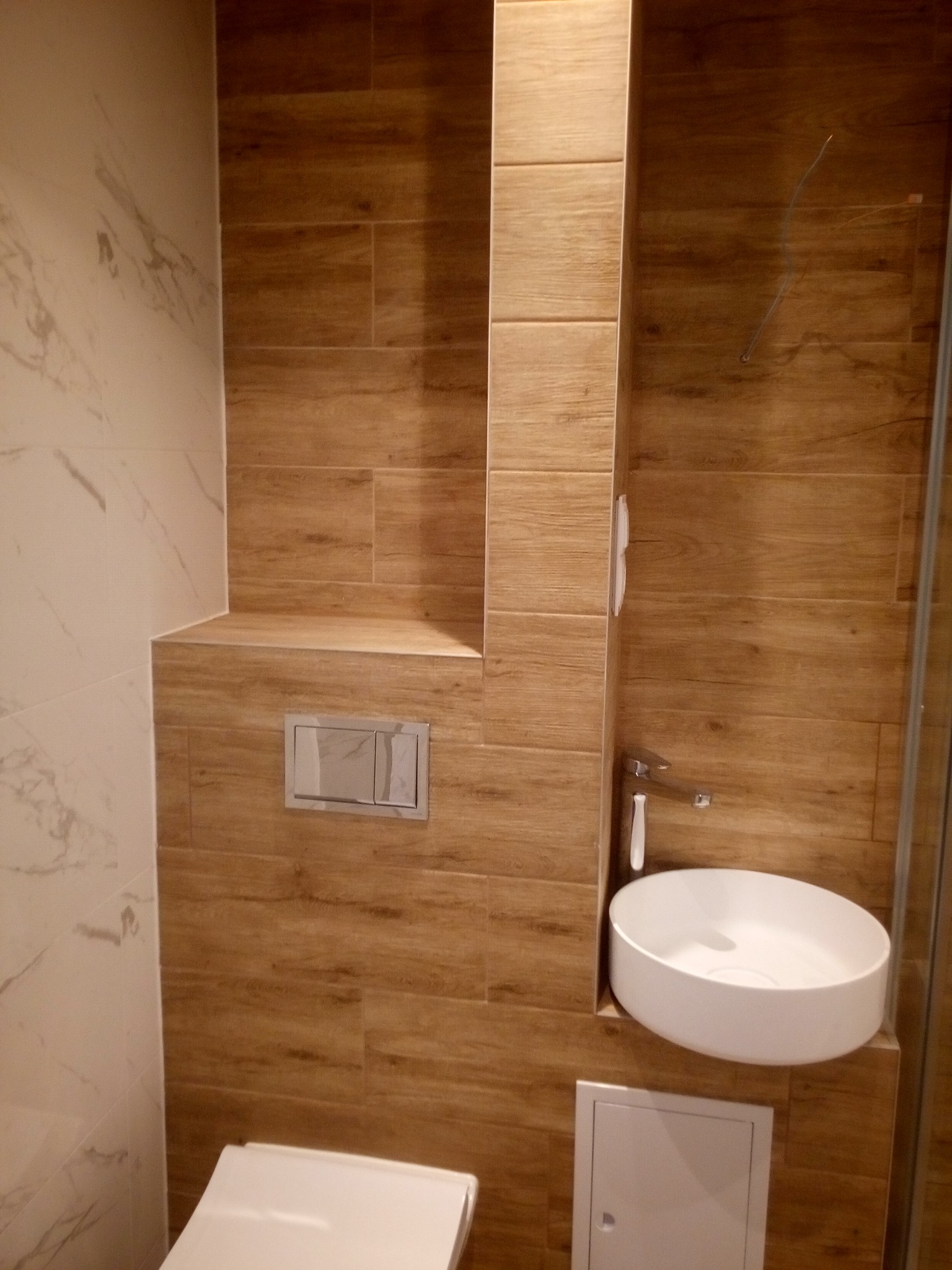 Nowoczesna łazienka z drewnopodobnymi płytkami, białą umywalką nablatową i podtynkową spłuczką WC, wykończona marmurową płytką.