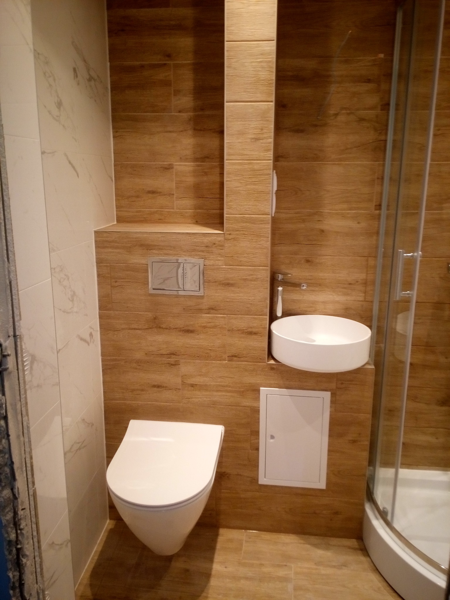 Nowoczesna łazienka z drewnopodobnymi płytkami, wiszącą miską WC, okrągłą umywalką nablatową i kabiną prysznicową z zaokrąglonymi drzwiami.