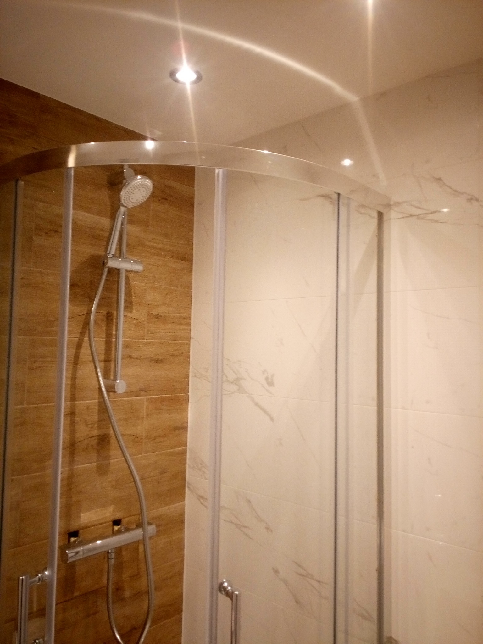 Kabina prysznicowa narożna z chromowanymi profilami i deszczownicą, wykończona płytkami imitującymi drewno i marmur, oświetlona punktowym światłem.