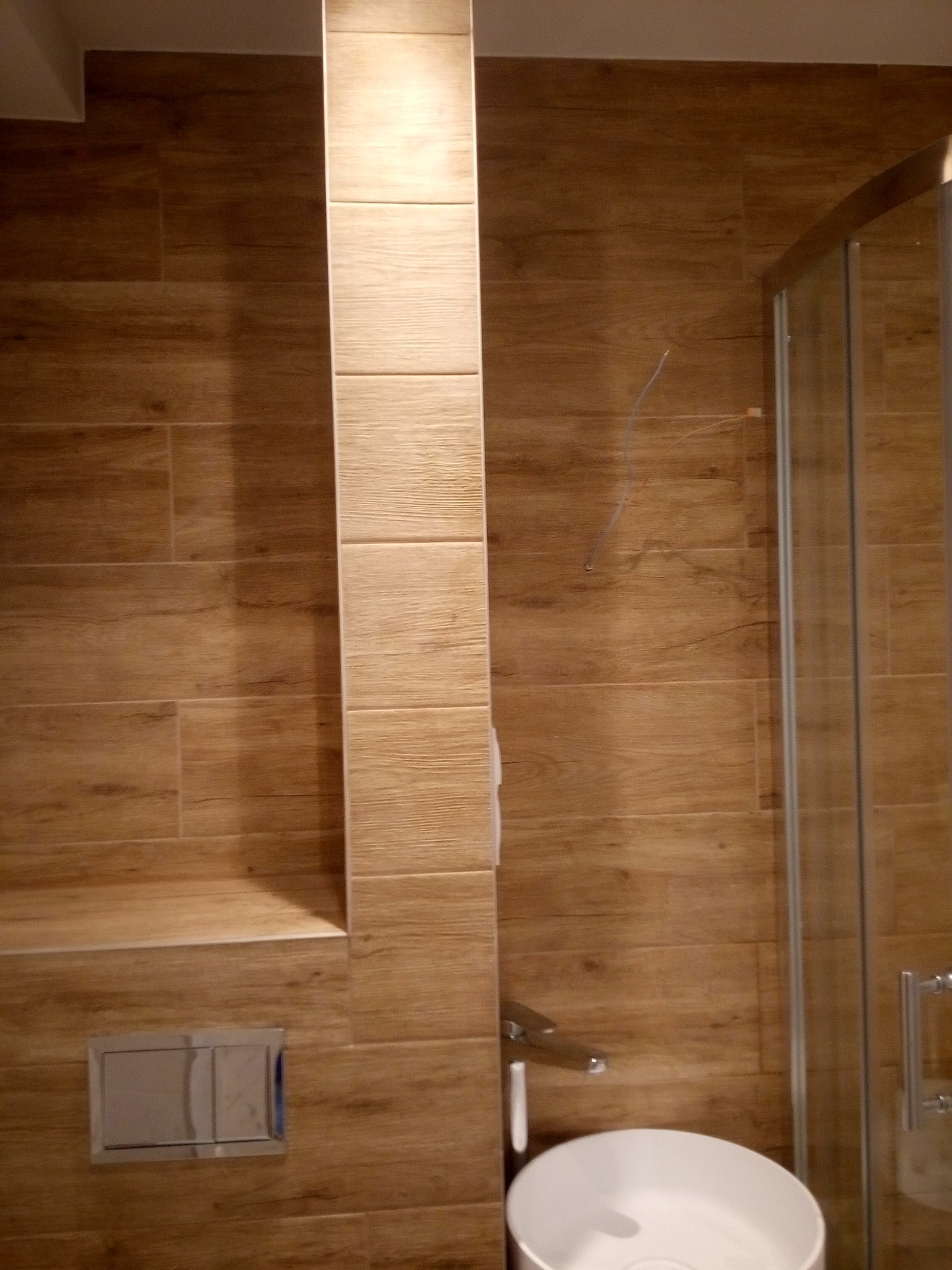 Wykończona łazienka z drewnopodobnymi płytkami, zabudową geberit i kabiną prysznicową z chromowanymi okuciami.