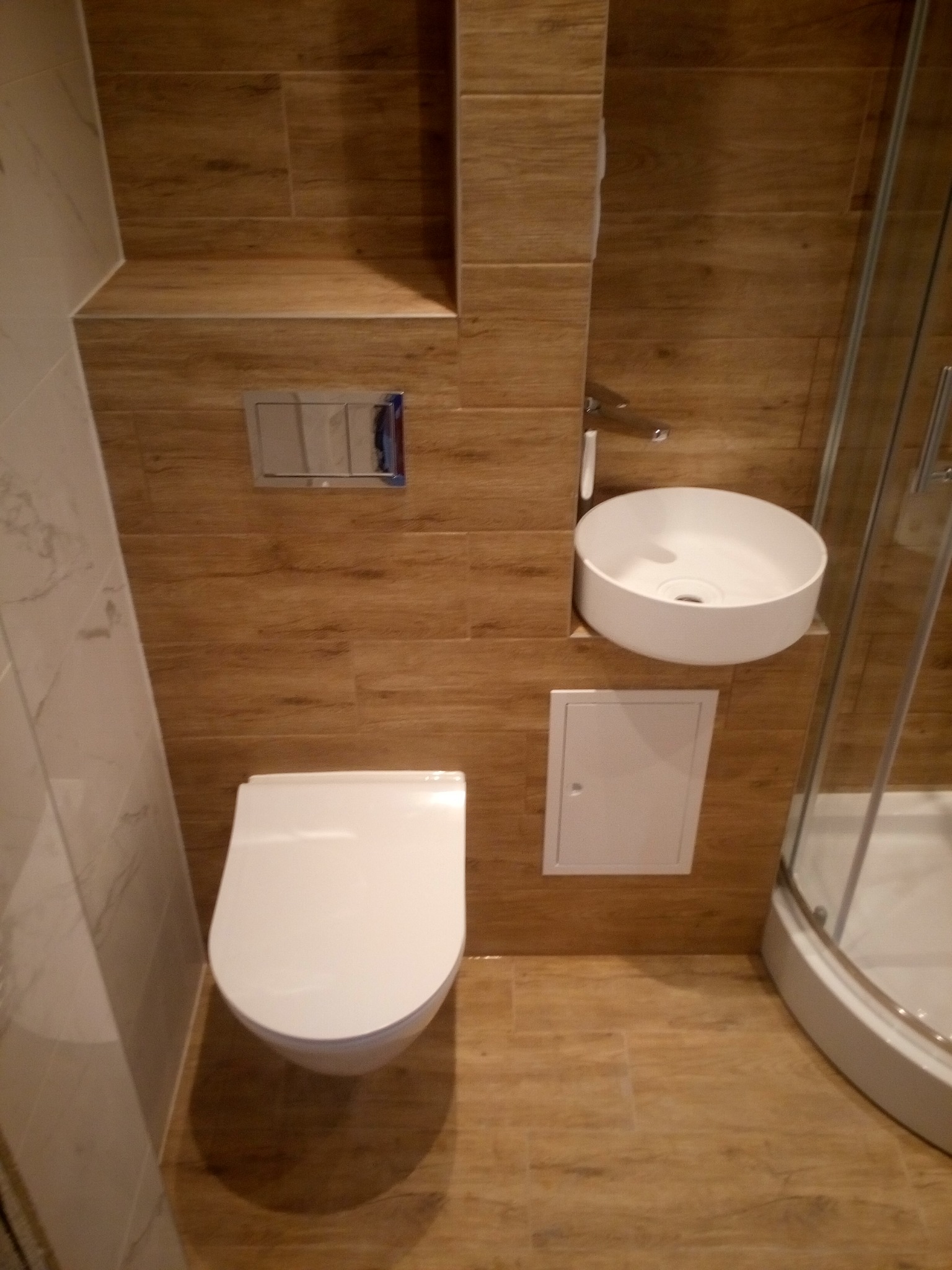 Nowoczesna łazienka z drewnopodobnymi płytkami, okrągłą umywalką nablatową i kabiną prysznicową z zaokrąglonym profilem.