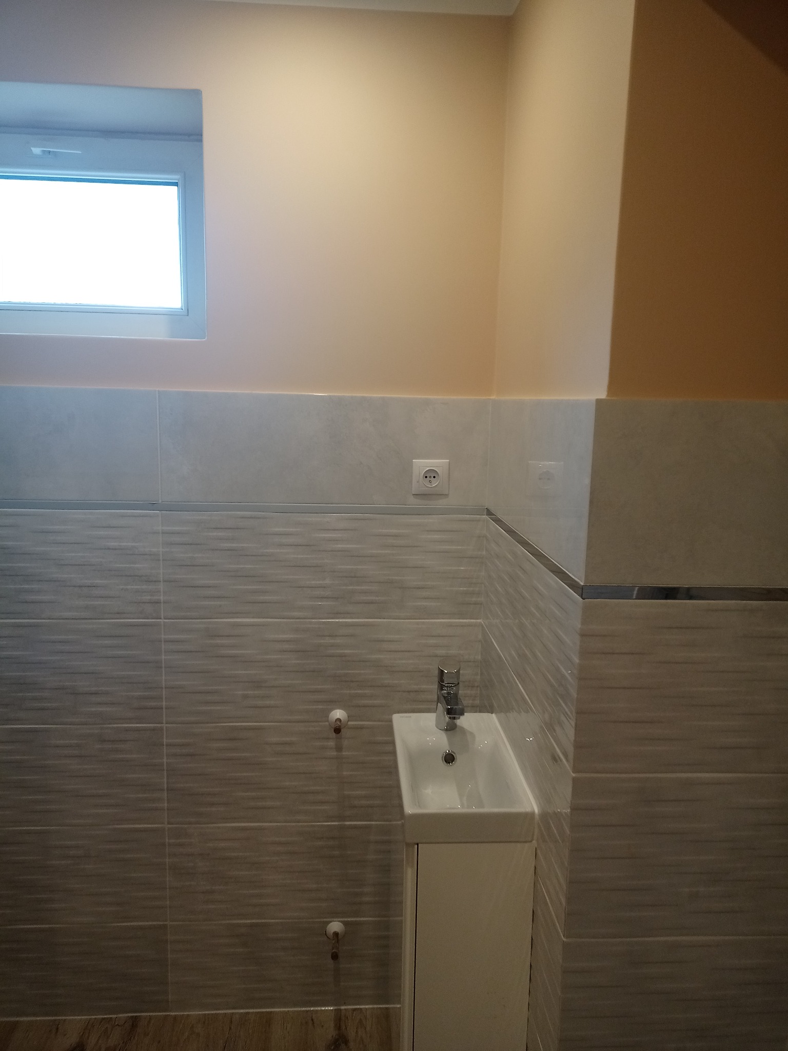 Nowoczesna łazienka po remoncie: umywalka z szafką w kolorze białym, szare płytki z poziomym wzorem, świeżo pomalowane ściany w odcieniach beżu, okno z białymi ramami.