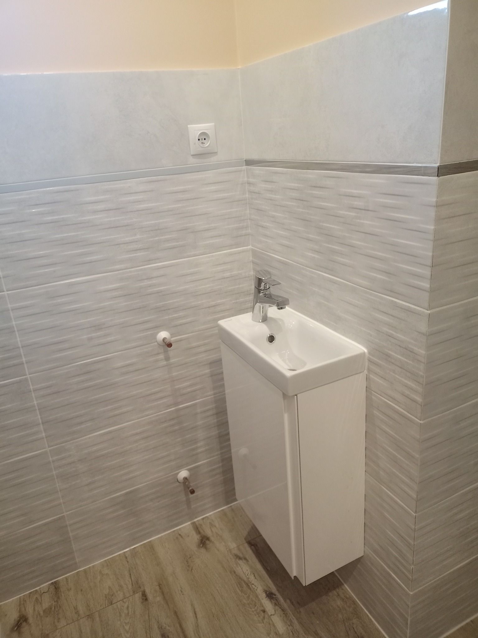 Nowoczesna, biała umywalka z szafką w łazience wyłożonej szarymi, poziomymi płytkami z delikatnym wzorem, drewnopodobna podłoga, widoczne przyłącza wodne i gniazdko elektryczne.
