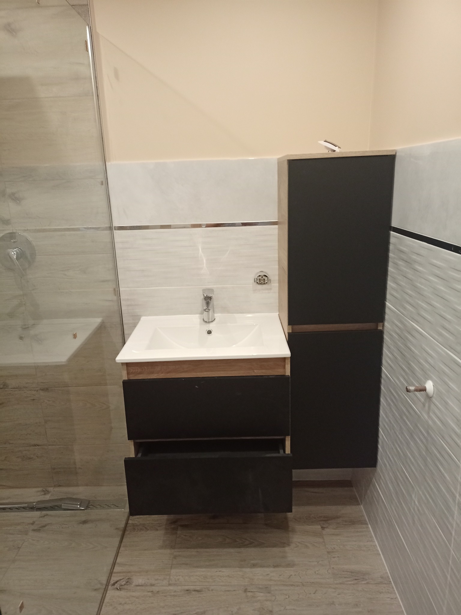 Nowoczesna łazienka z umywalką nablatową, szafką z szufladami z otwartą jedną szufladą, wysoką szafką boczną i kabiną prysznicową w tle, wykończona płytkami w odcieniach szarości i beżu.