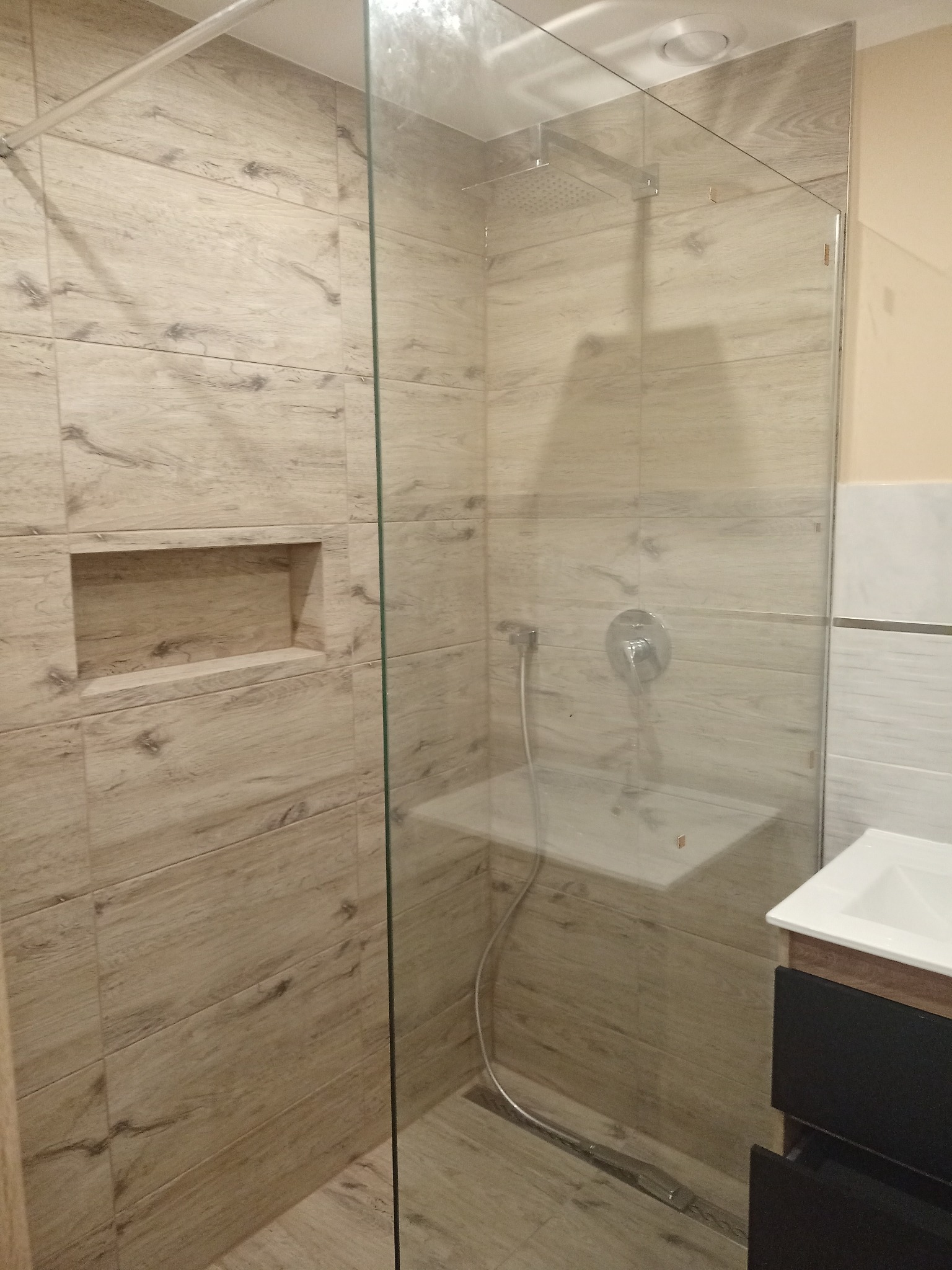 Nowoczesna kabina prysznicowa z transparentną szybą, wykończona płytkami imitującymi drewno, z wnęką w ścianie i czarną szafką pod umywalką.